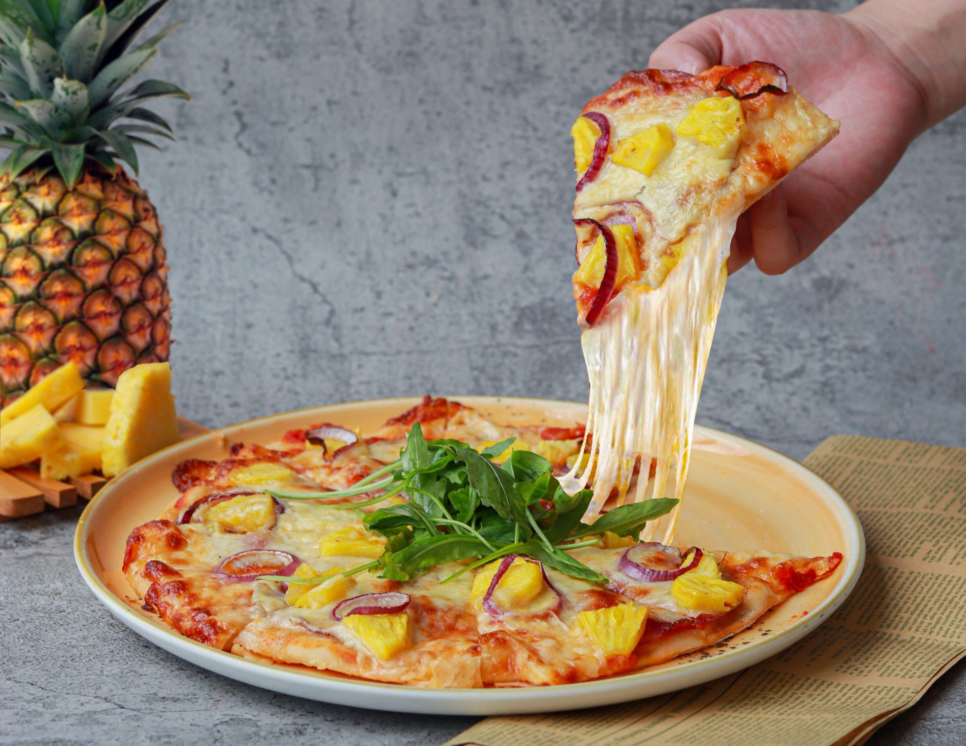 Hand pulls a pineapple pizza slice with stretching cheese; a whole pineapple is in the background.