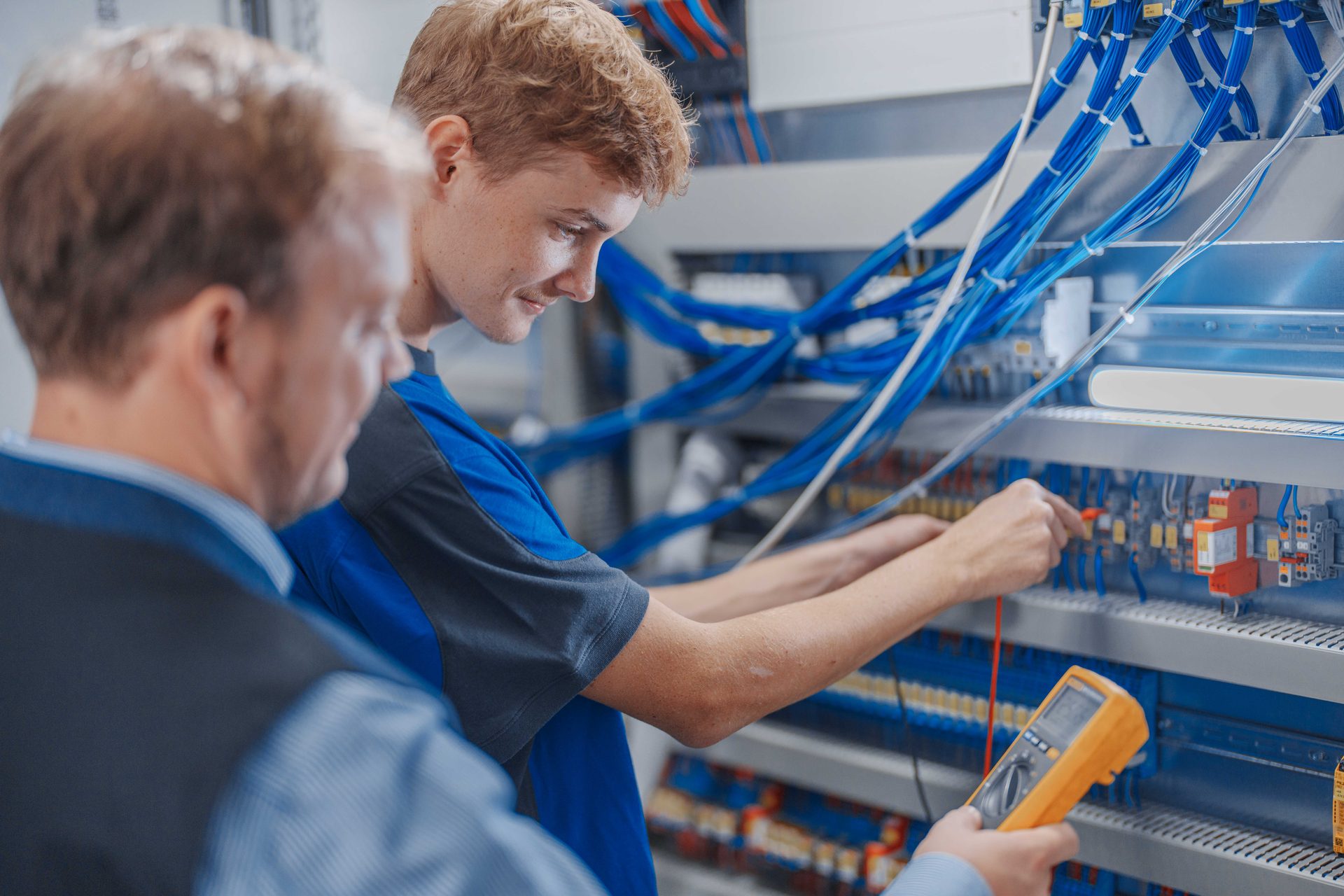 Two electricians troubleshoot an electrical panel with a multimeter.