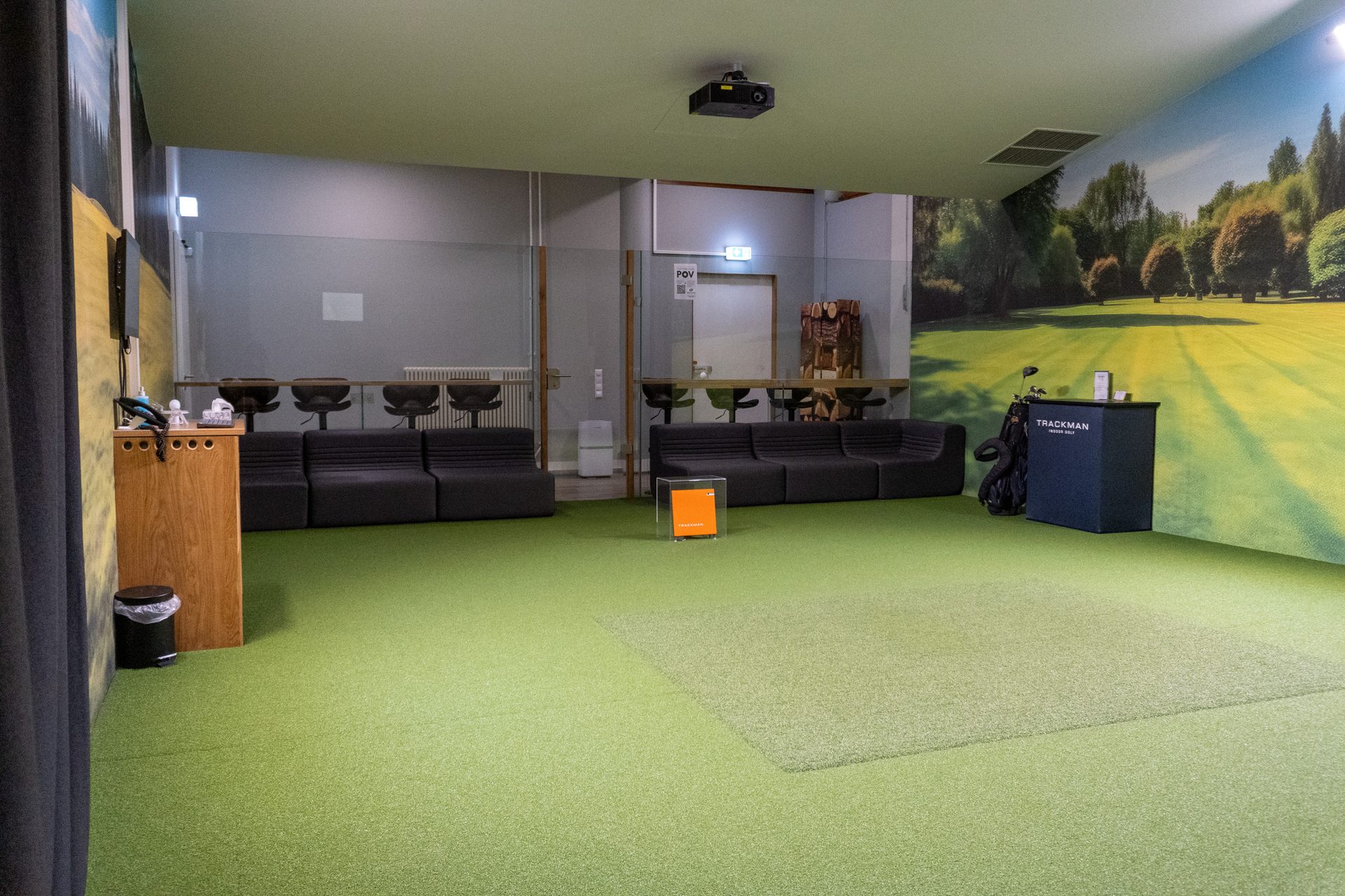 Indoor golf simulator room with green turf, black sofas, Trackman stand, projector, and nature murals.