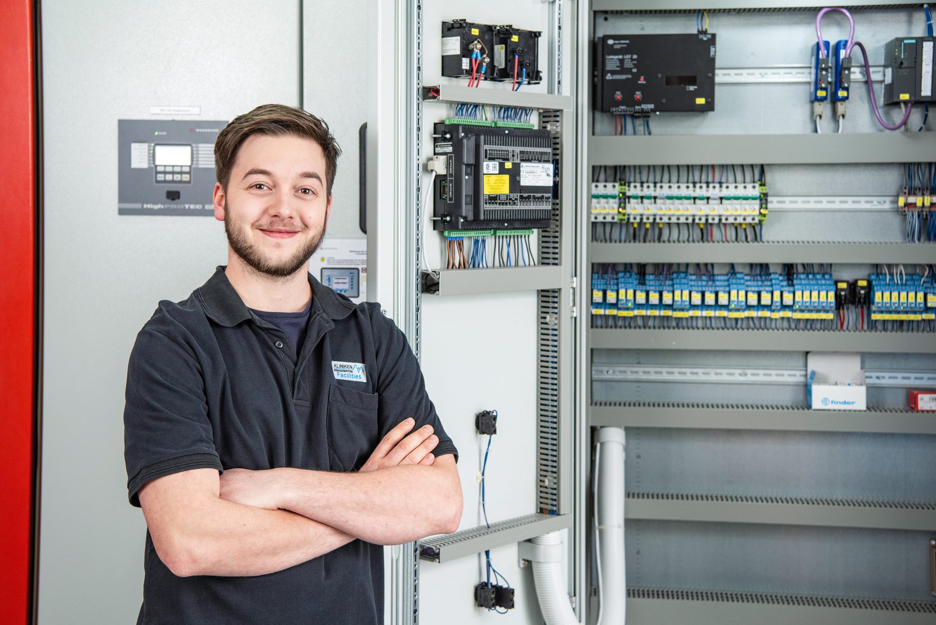 Smiling man stands by an open electrical control cabinet, showing wires and components.