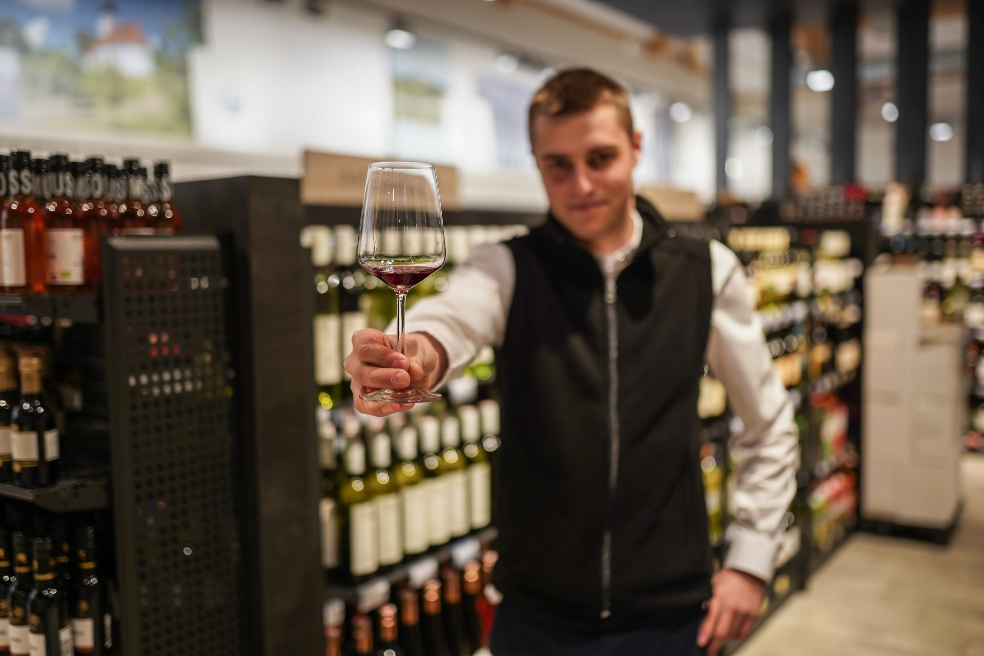 Man holding a glass of red wine towards the viewer in a store.