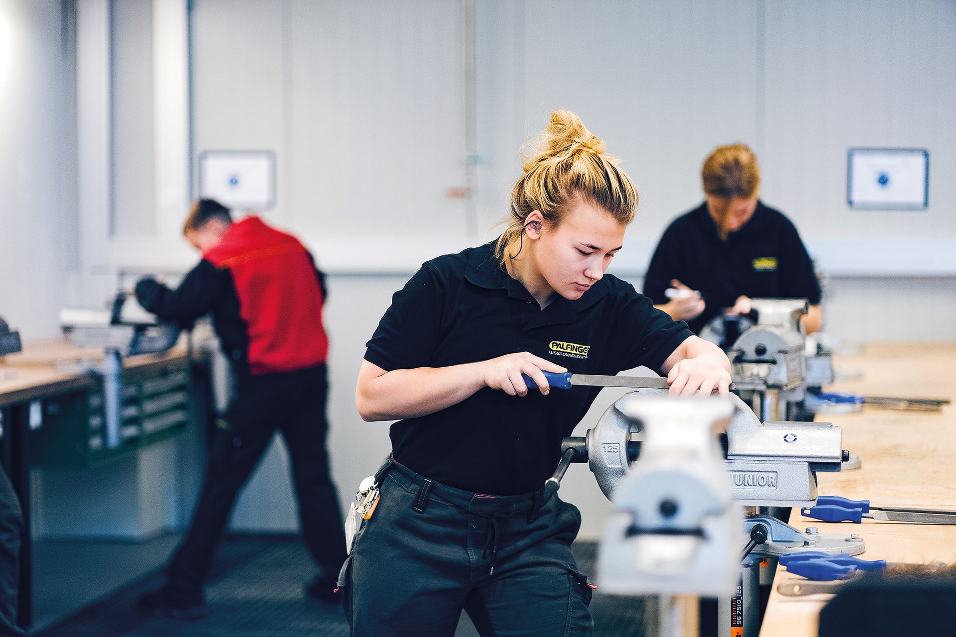 Young woman filing metal at a workshop bench. Two others work blurred in the background.
