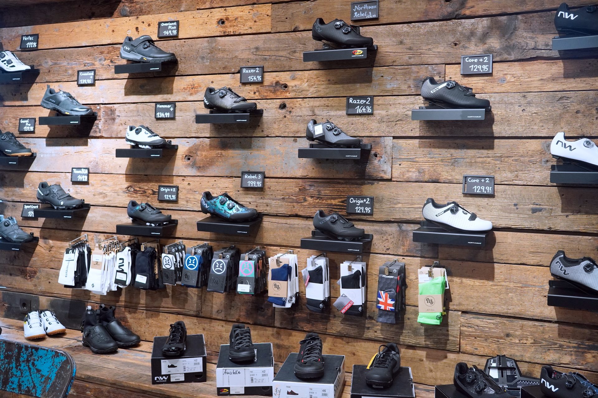 A wooden wall display showcasing various models of cycling shoes and socks, with prices visible.