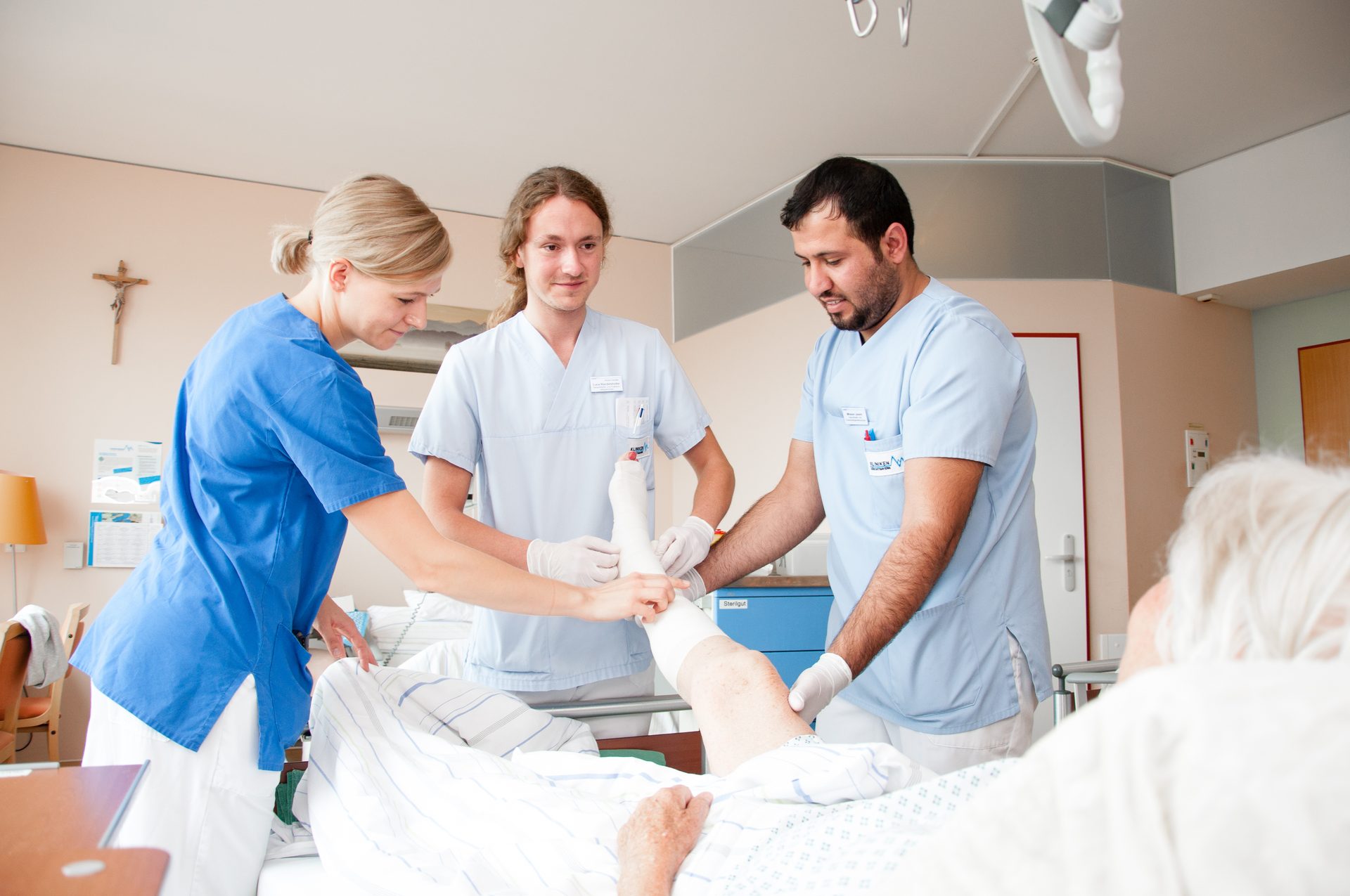 Three medical staff apply a cast to a patient's leg in a hospital room.