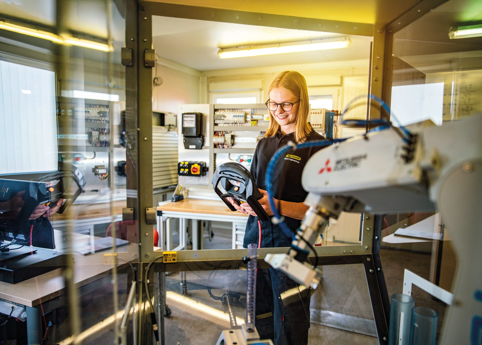 Smiling woman in glasses holds a controller in a tech lab with a robot arm.