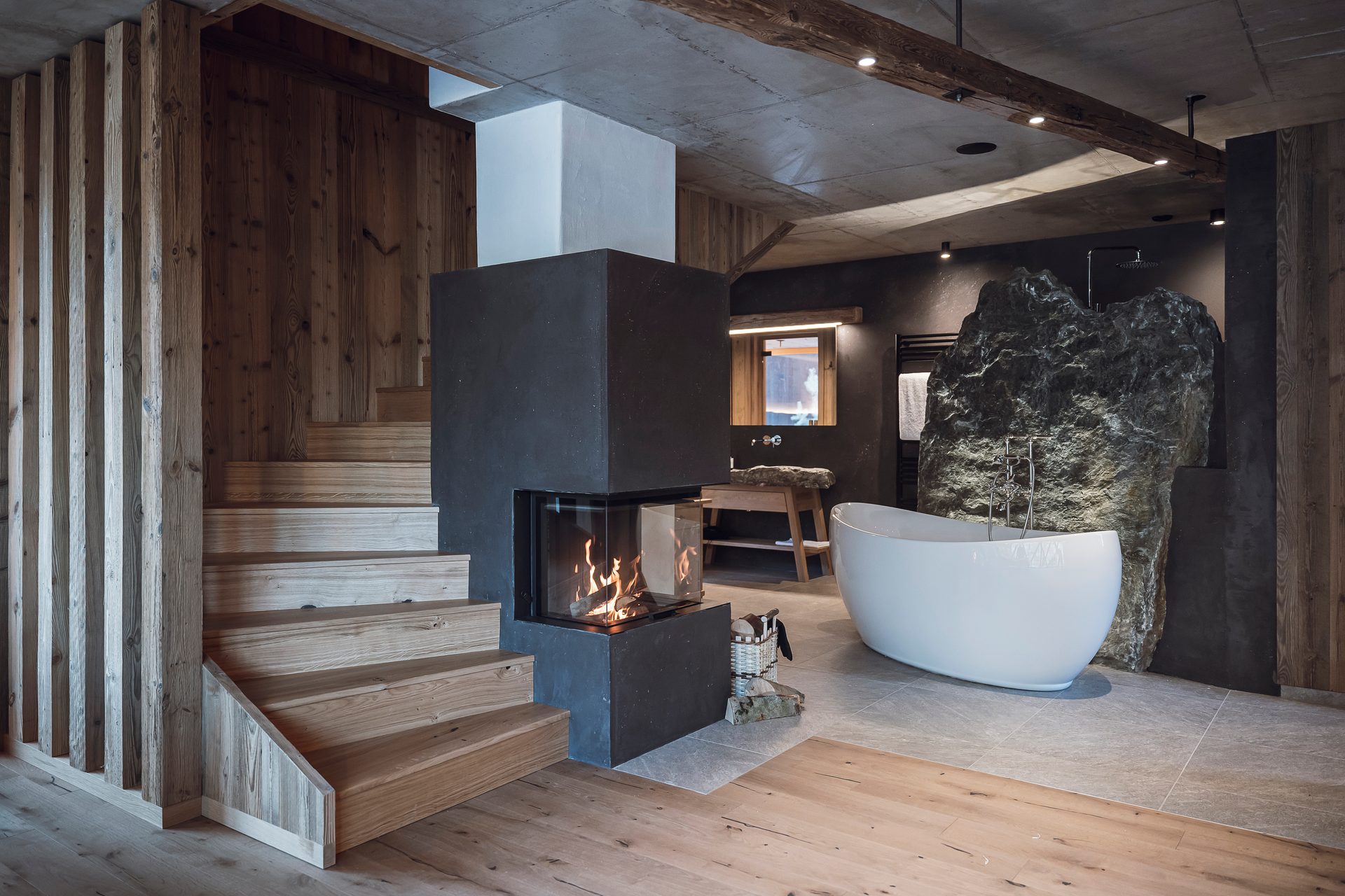 Rustic modern bathroom with a freestanding tub, rock wall, and wood staircase next to a fireplace.
