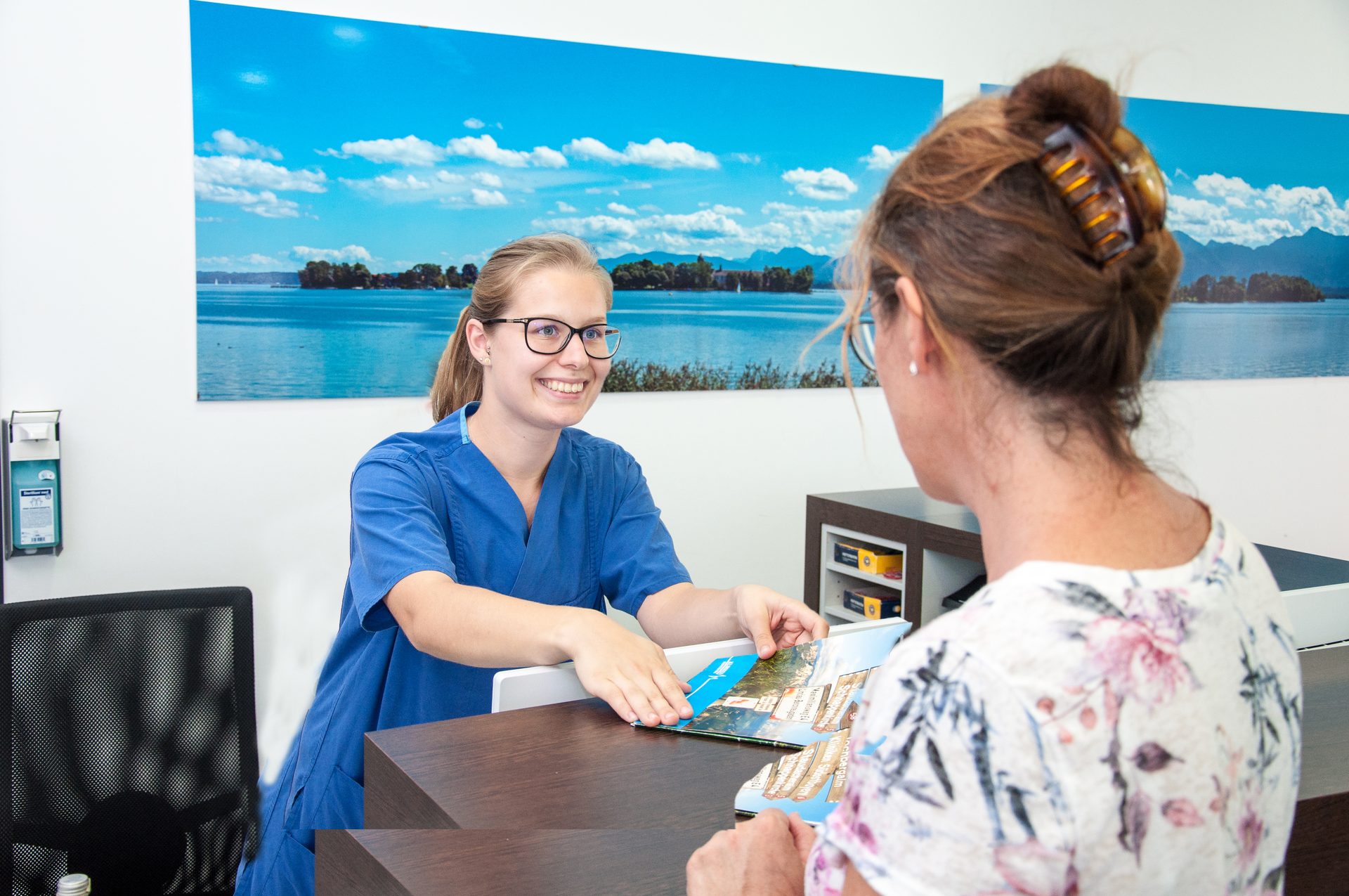 A smiling woman in blue scrubs shows a brochure to a customer at a reception desk.
