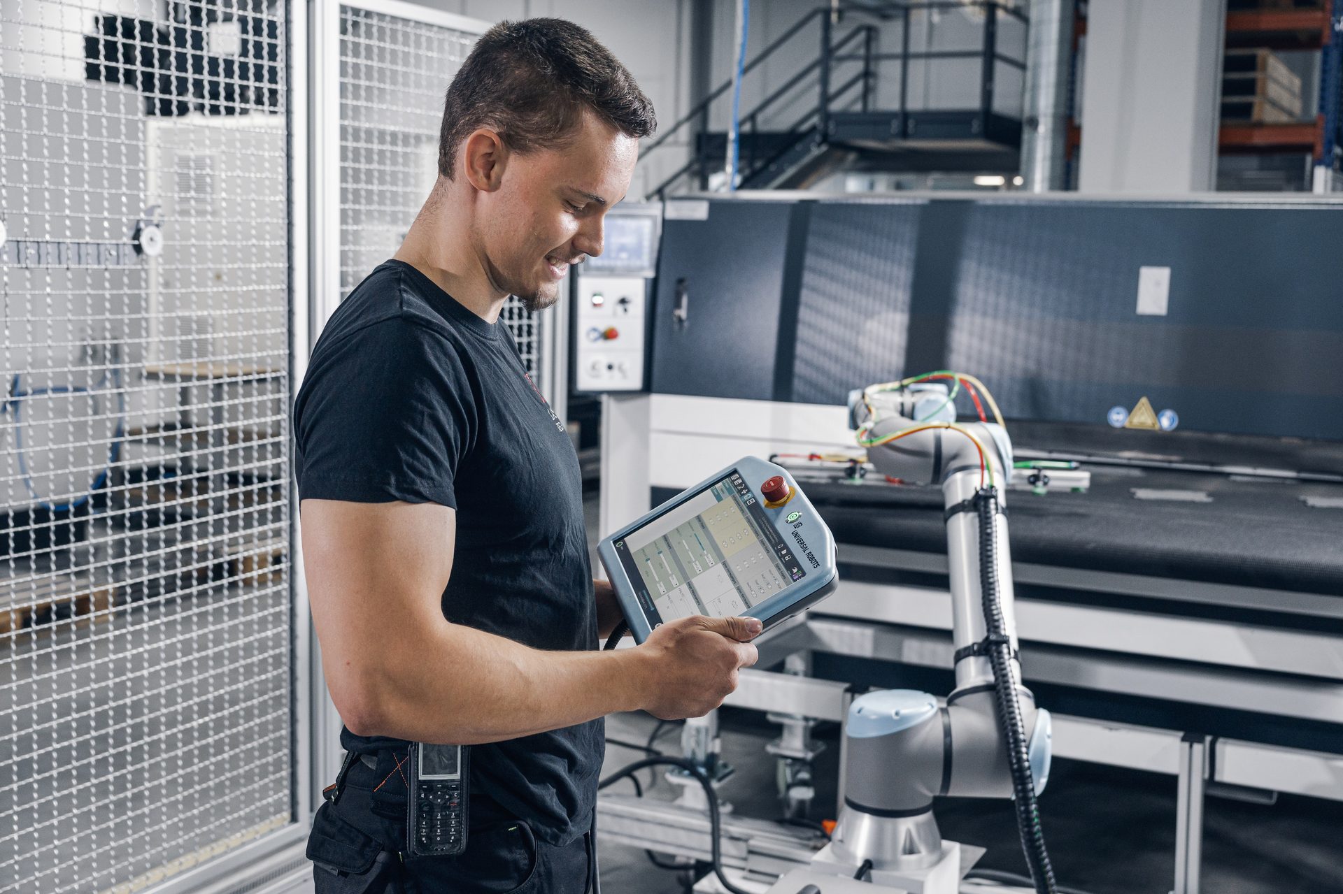 Man smiling, operating a robotic arm with a tablet in a factory setting.
