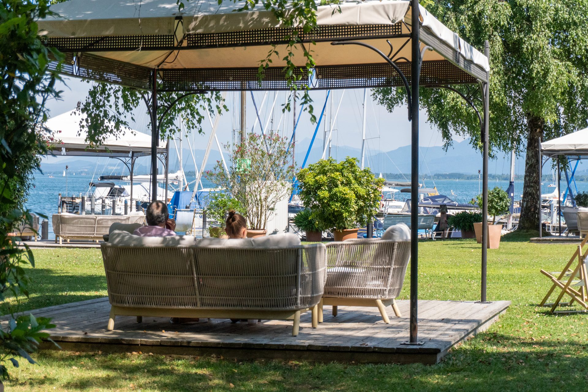 People under a lakeside gazebo overlooking boats and distant mountains.