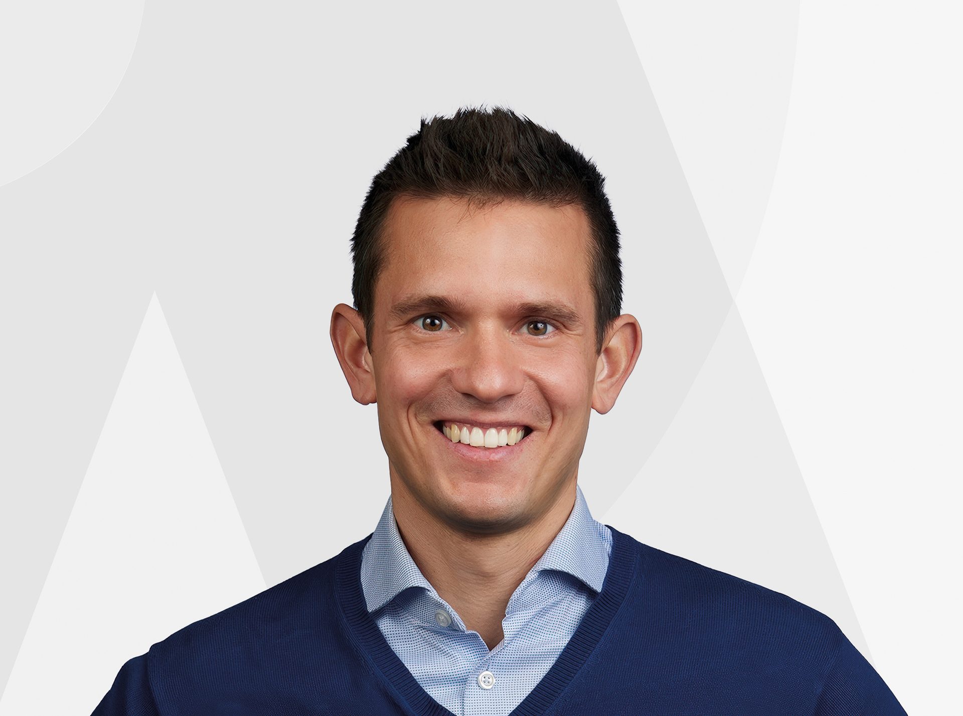 A smiling man in a blue V-neck sweater over a patterned light blue shirt.