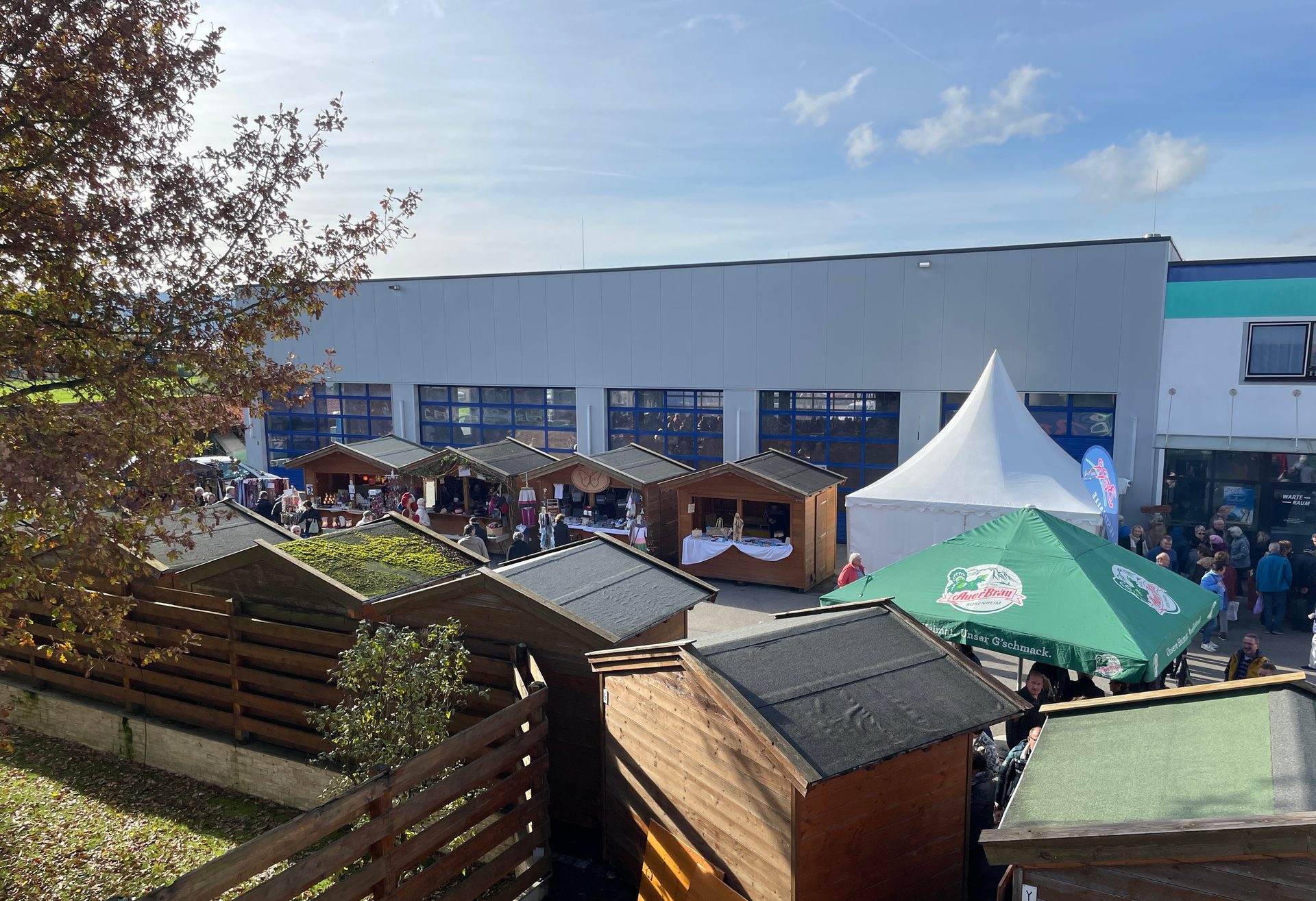 An outdoor market scene with wooden stalls, a large white tent, a green umbrella, and people browsing.