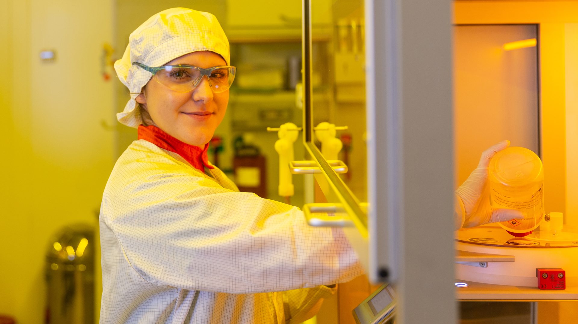 Smiling scientist in protective gear operates equipment in a yellow-lit lab.