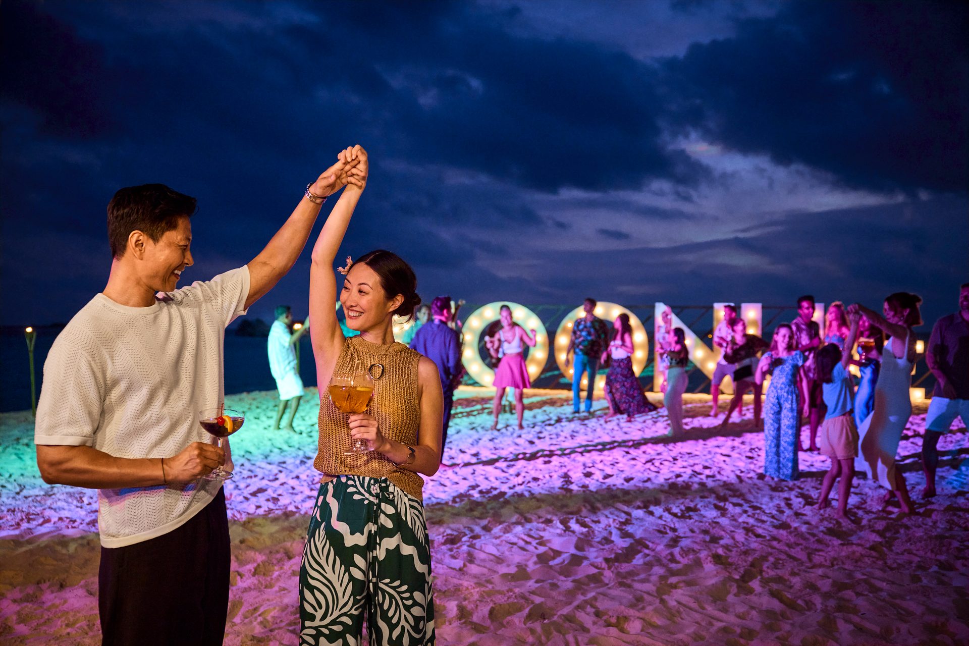 Couple and friends dance at a colorful night beach party with glowing letters.