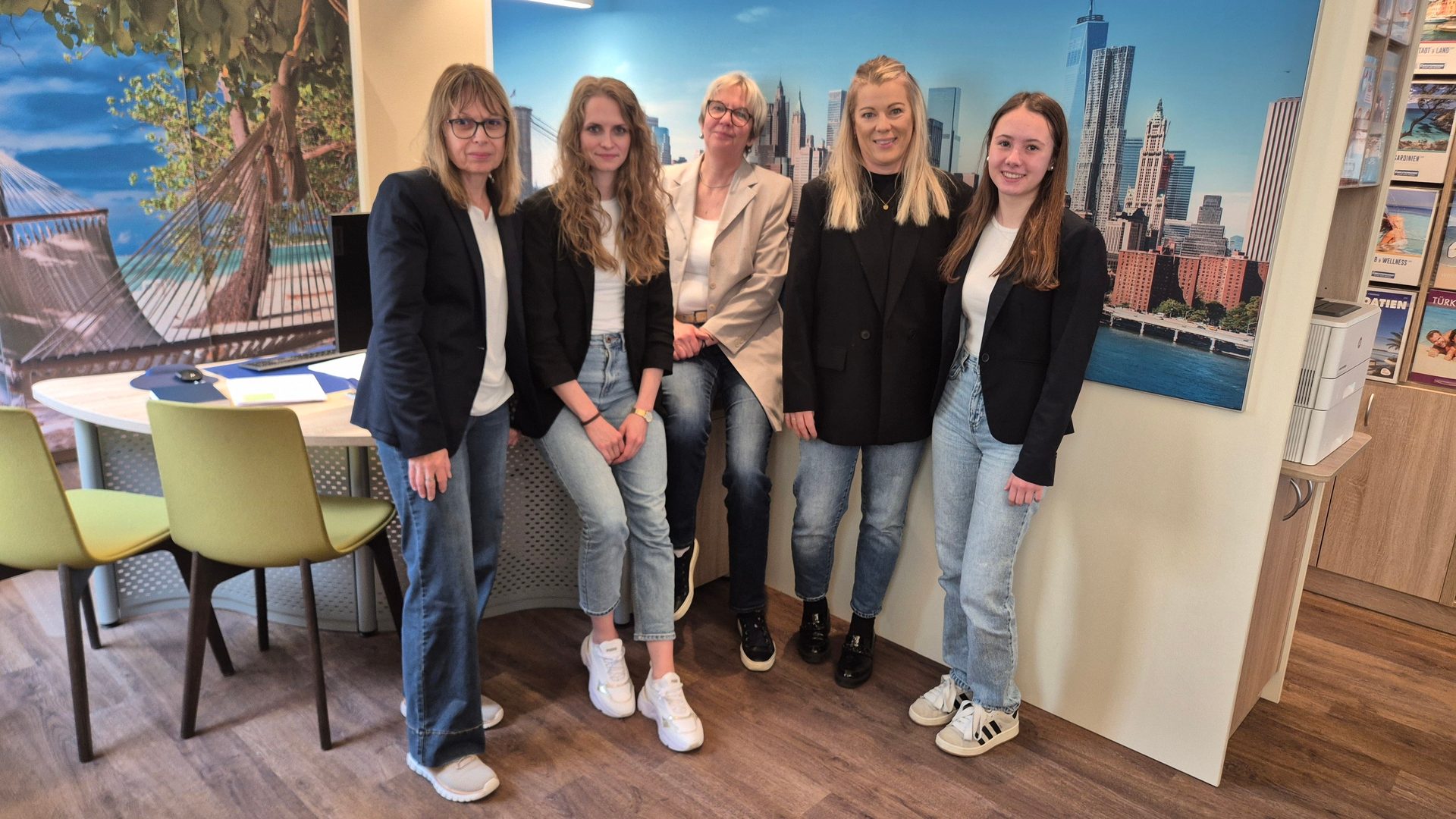 Five women in blazers and jeans stand smiling in an office with travel-themed murals.
