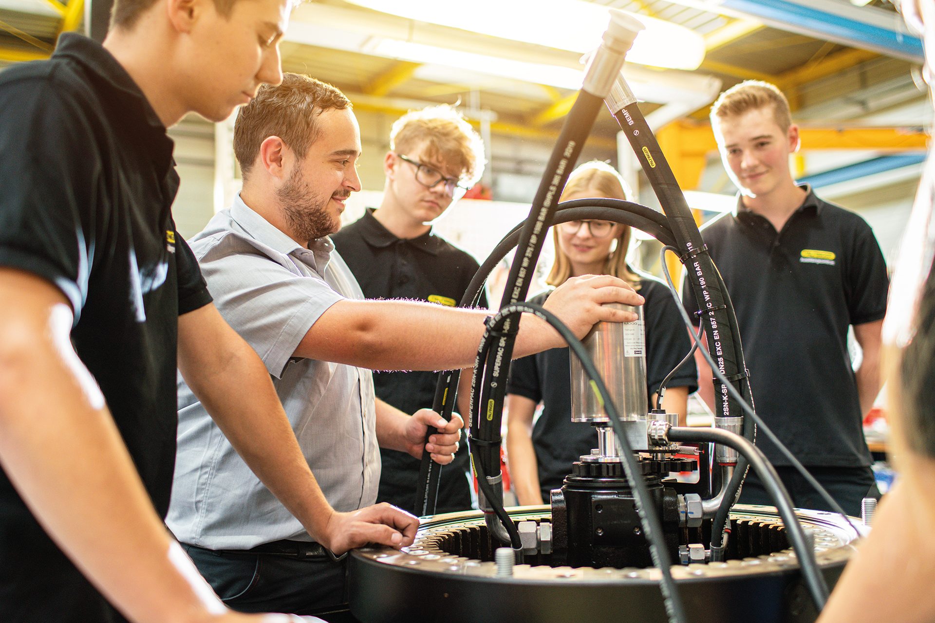 An instructor shows industrial machinery with hoses and gears to four apprentices in a workshop.