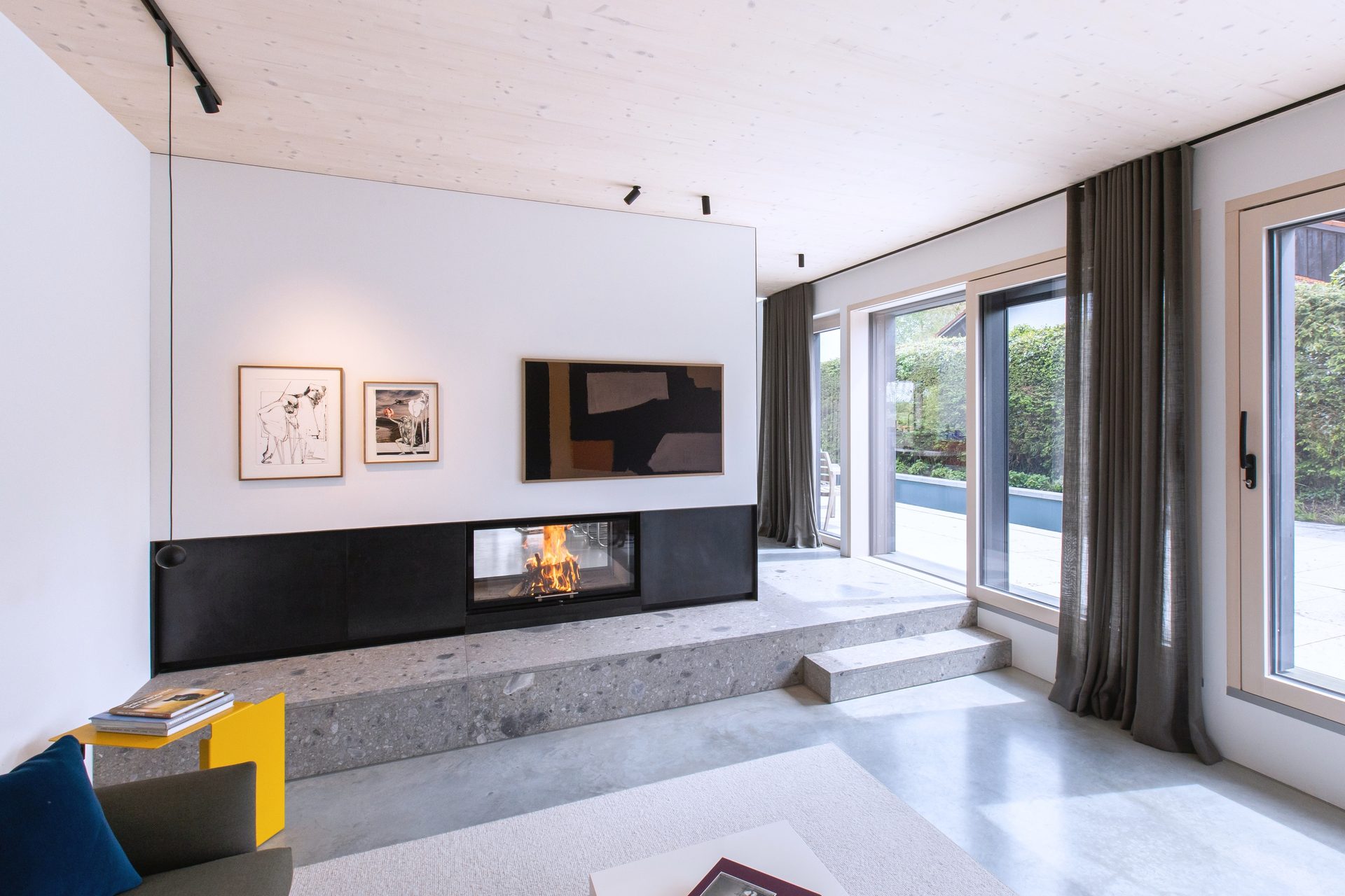 Modern living room with a fireplace, abstract art, large windows, wooden ceiling, and concrete steps.