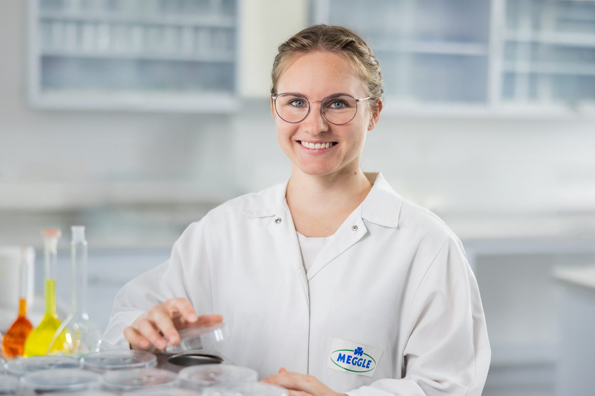 Smiling female scientist in MEGGLE lab coat holds petri dish in lab.