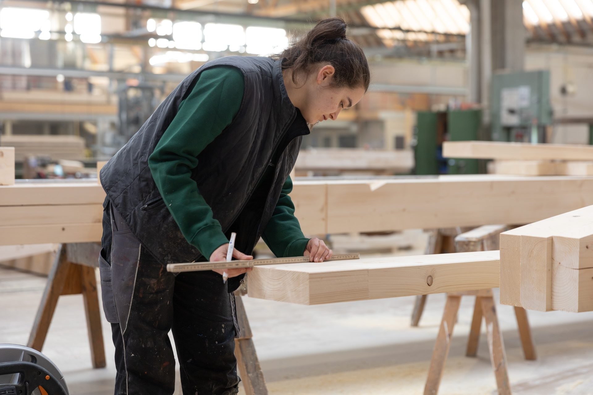 Young woman in workwear measuring a wooden beam with a ruler in a carpentry workshop.