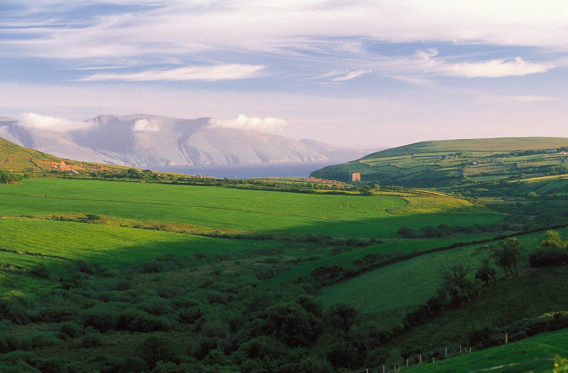 Vast green Irish landscape with rolling hills, a calm bay, distant mountains, and an old stone building.