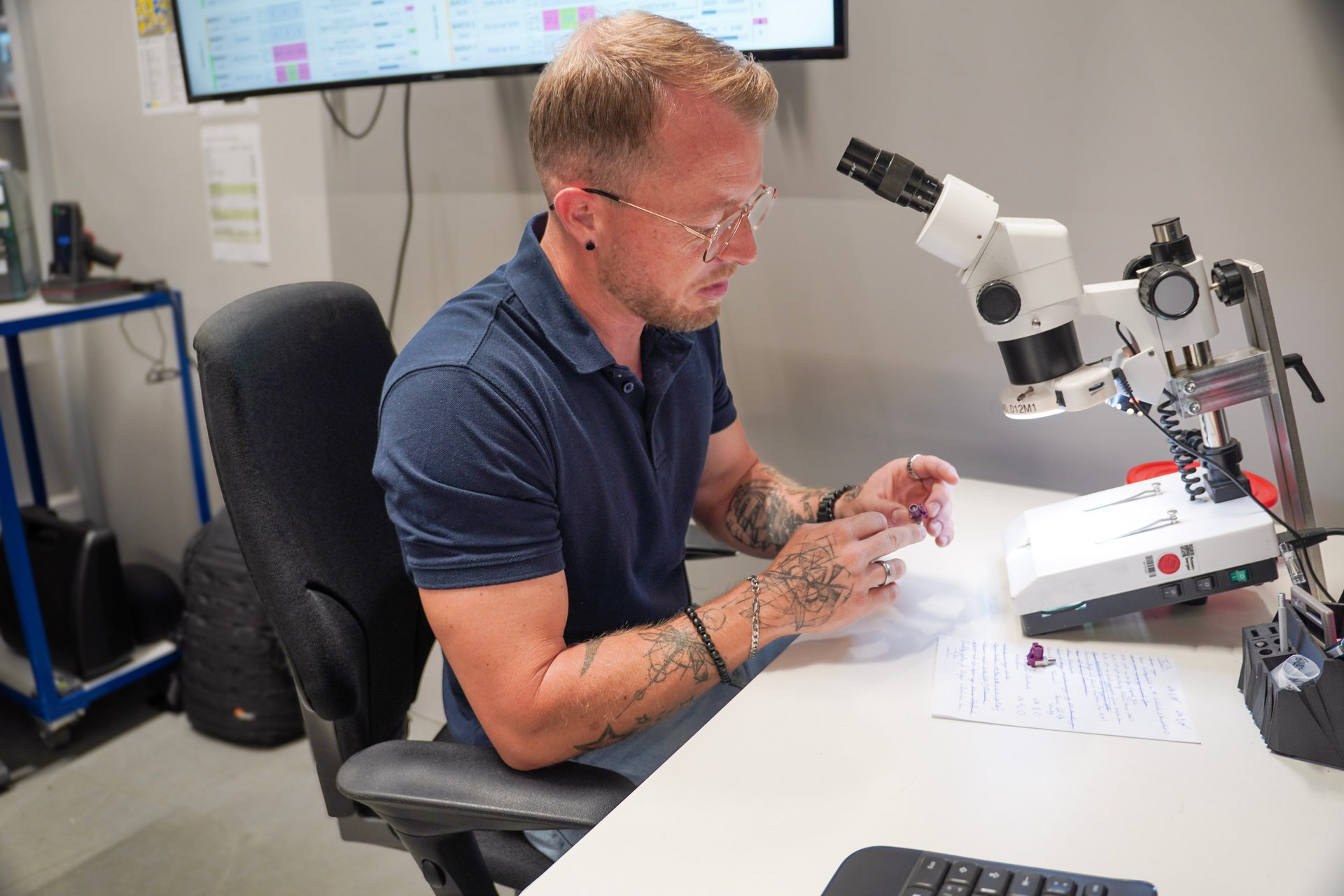 Man with glasses and tattooed arms examining small components at a desk with a microscope.