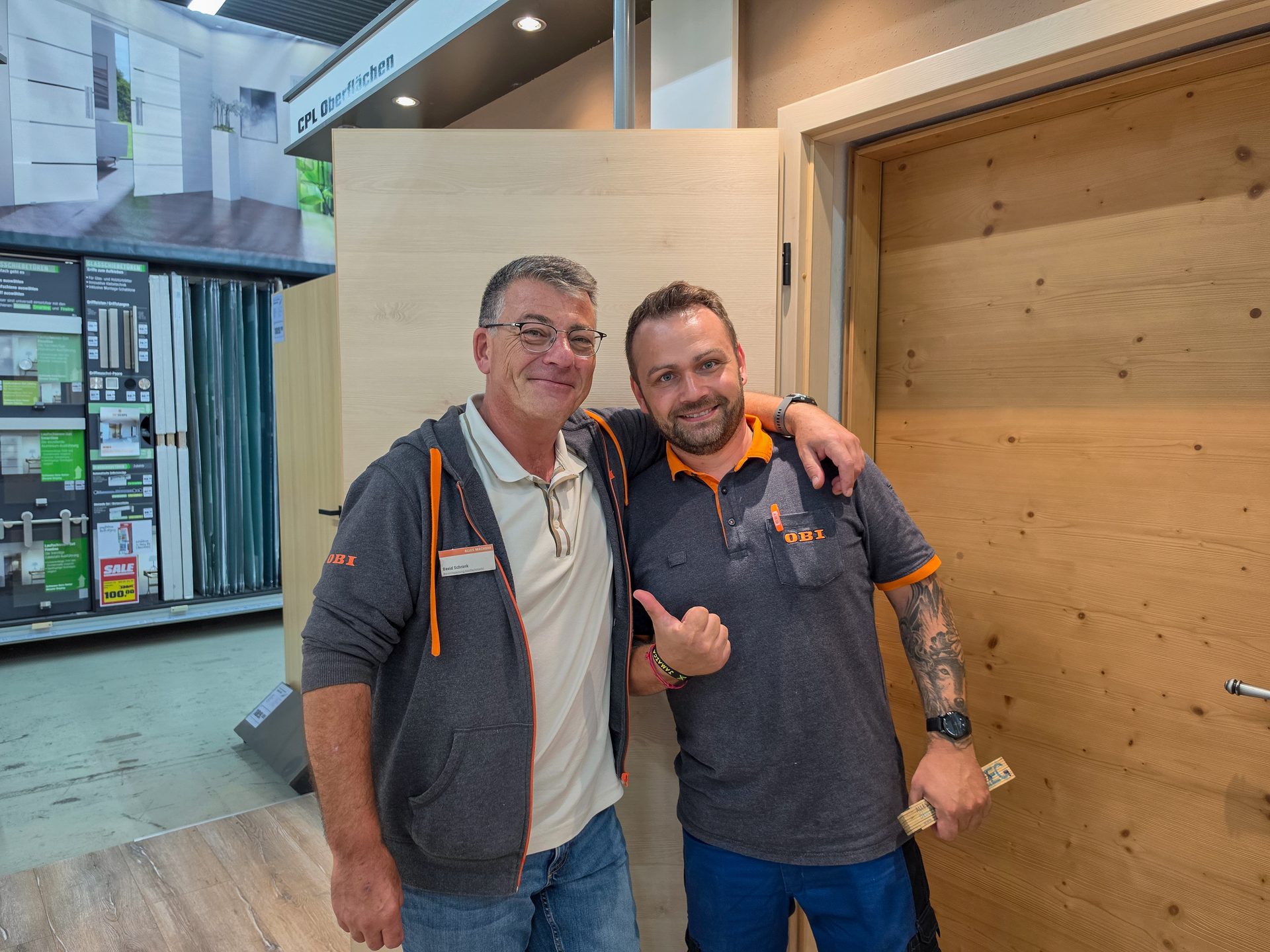 Two smiling men, likely OBI store employees, stand side-by-side in a home improvement store.