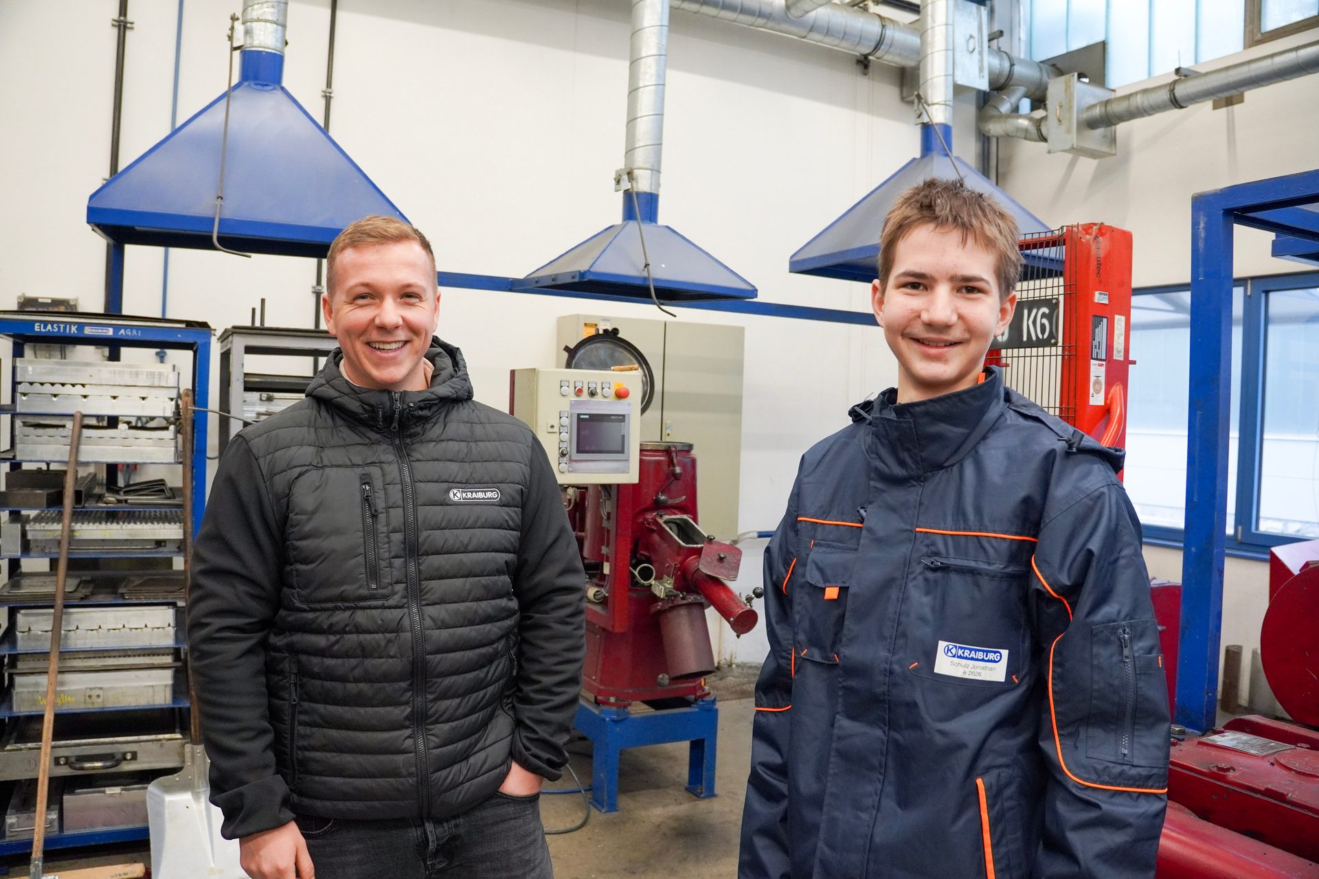 Two smiling men, one in a Kraiburg jacket, stand in a workshop with industrial machinery.