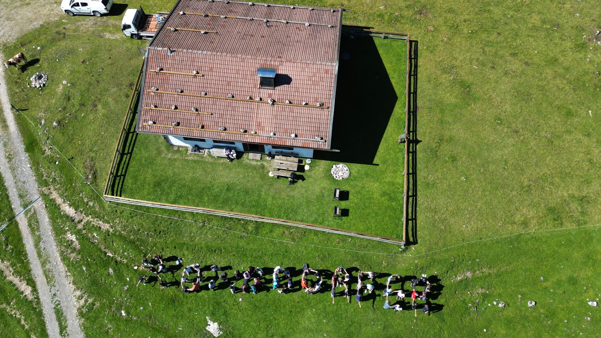 Aerial: Mountain hut, green field. People spell "SCHILCHER" on the grass.