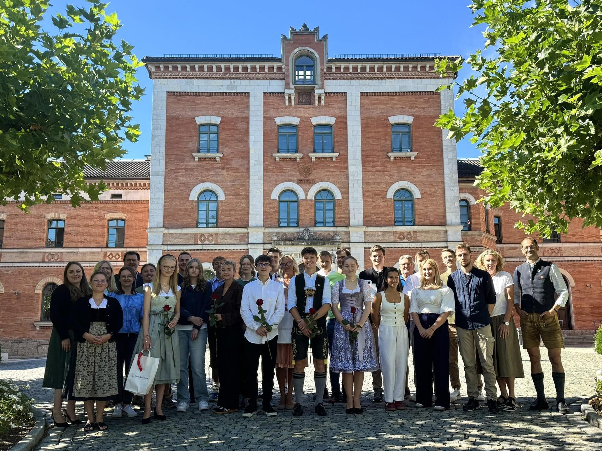 A group of people, some in traditional Bavarian attire, holding roses in front of a large brick building.