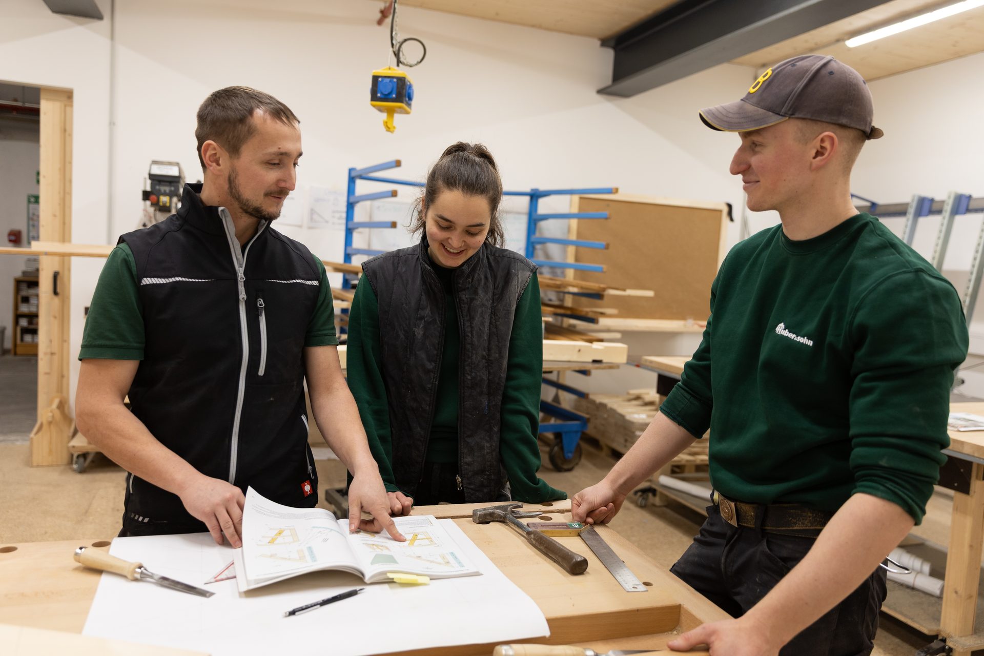 Three carpenters discuss plans in a woodworking workshop, pointing at a book.