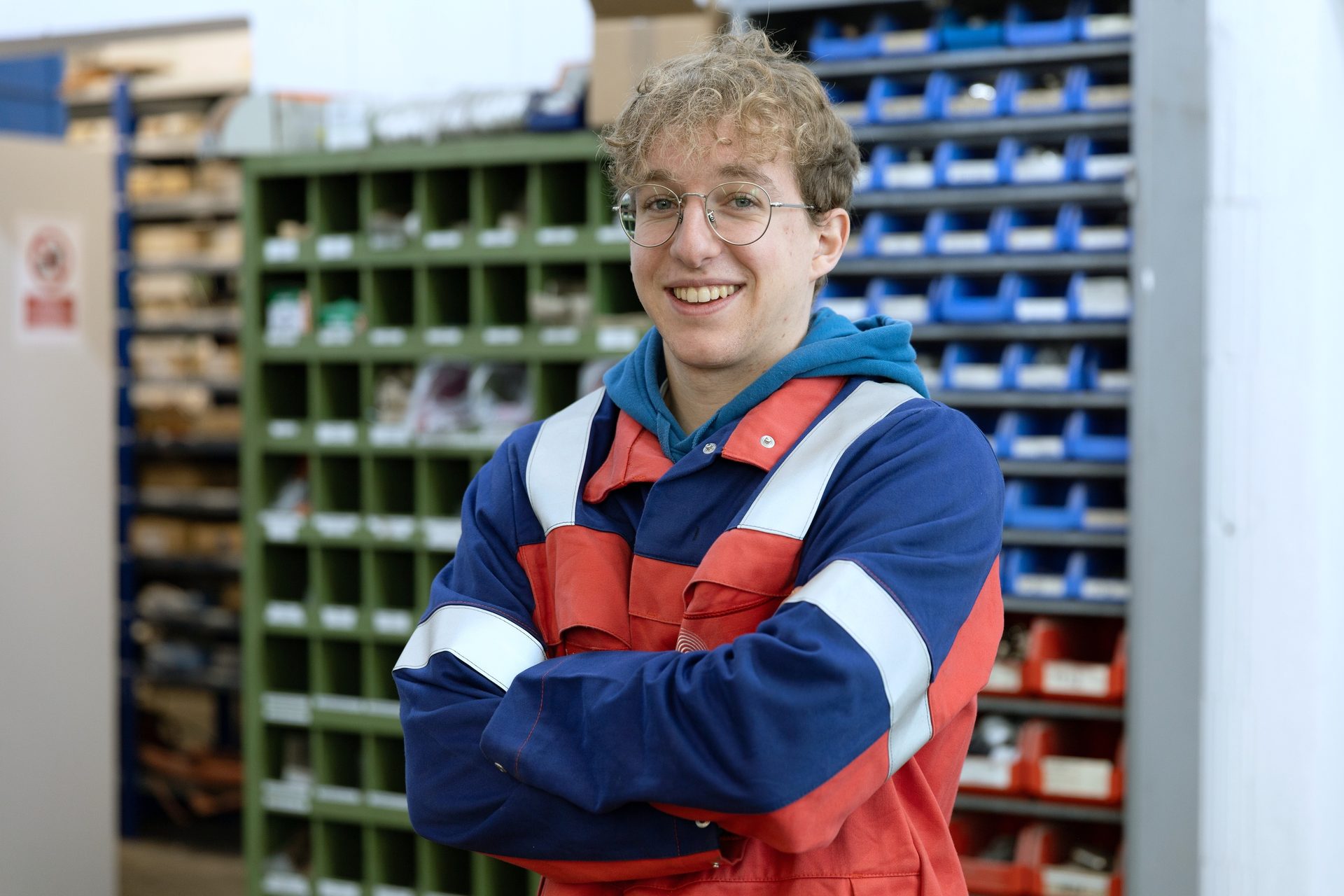 Smiling young man in a work jacket with crossed arms in a parts storage area.