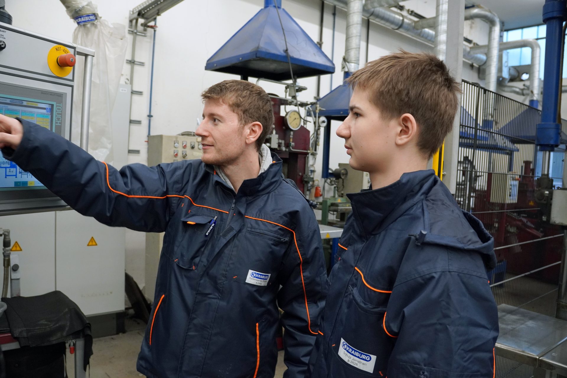 Two men in Kraiburg work jackets, one pointing at a touch screen control panel in a workshop.