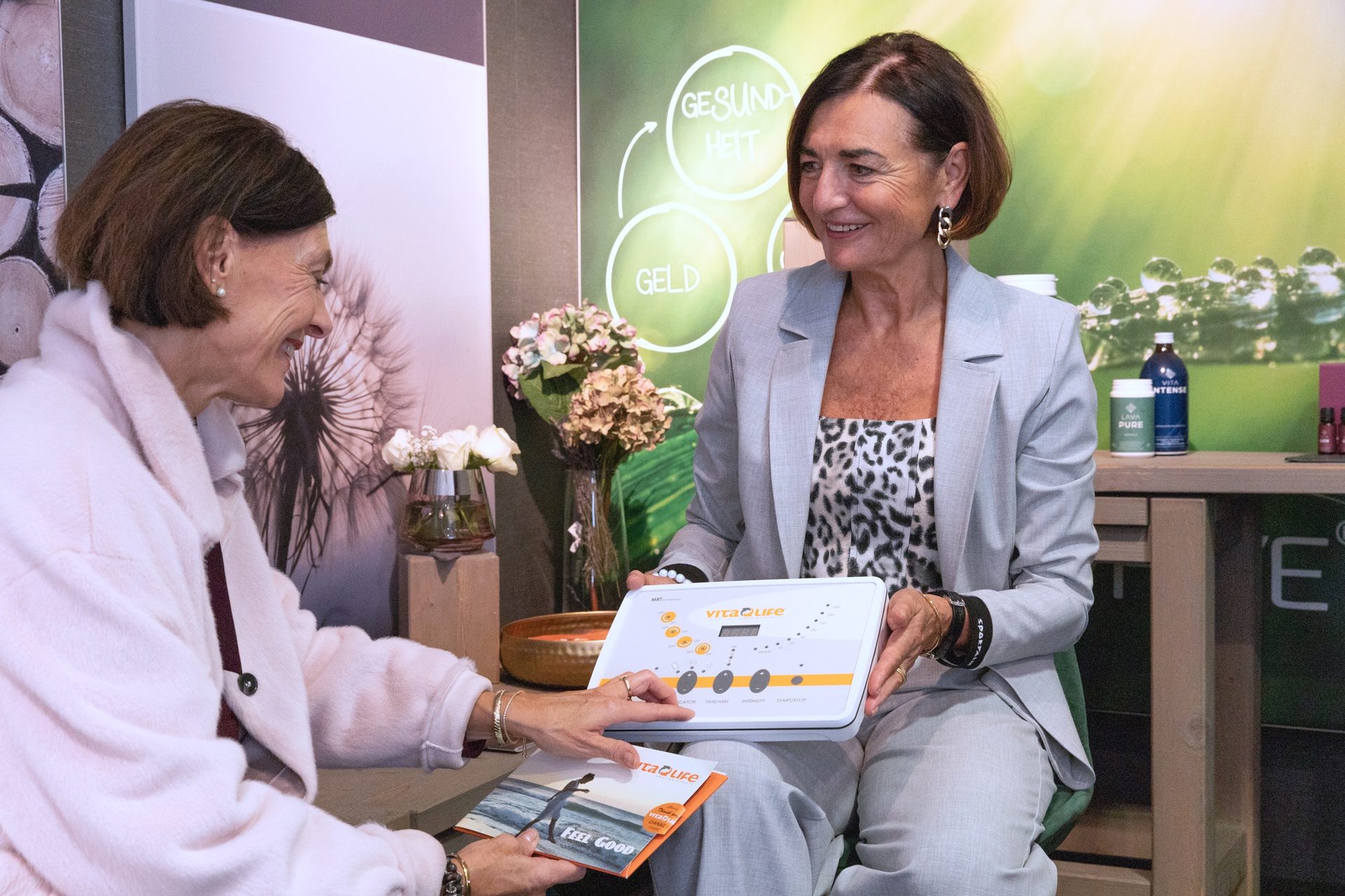 Two women at a wellness event; one presents a 'VITAQLIFE' device, the other holds a brochure.