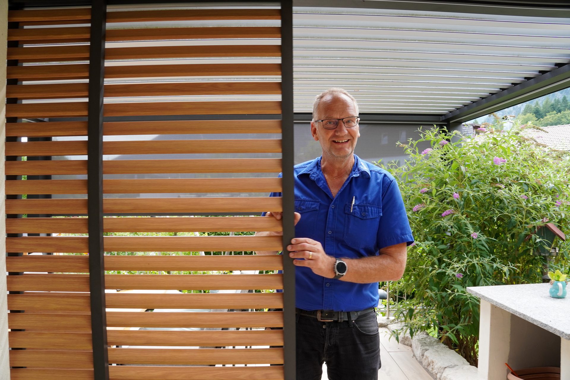A smiling man in glasses and a blue shirt stands behind a wooden slatted screen on a patio.