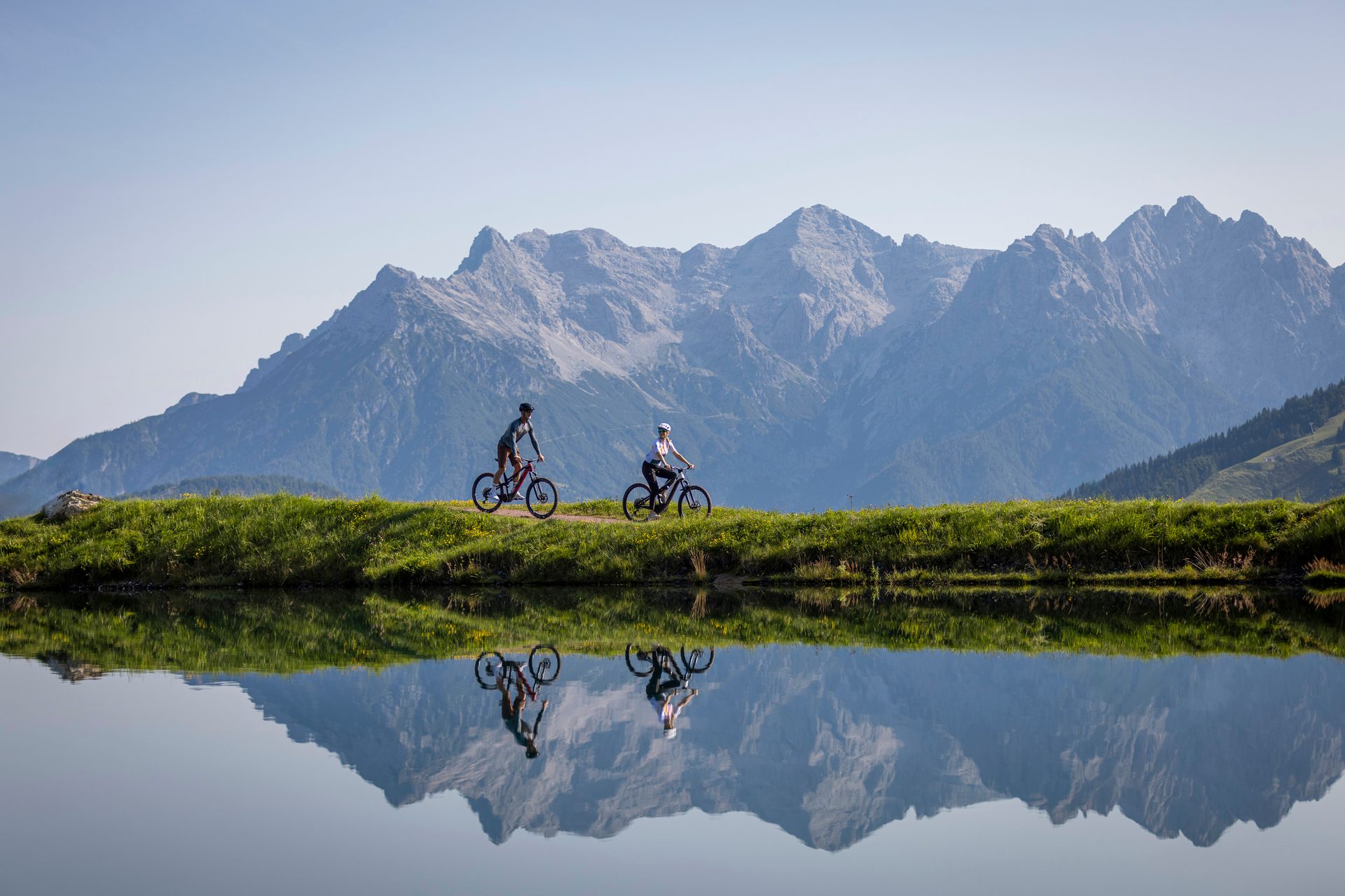 Two mountain bikers on a trail next to a lake reflecting mountains.