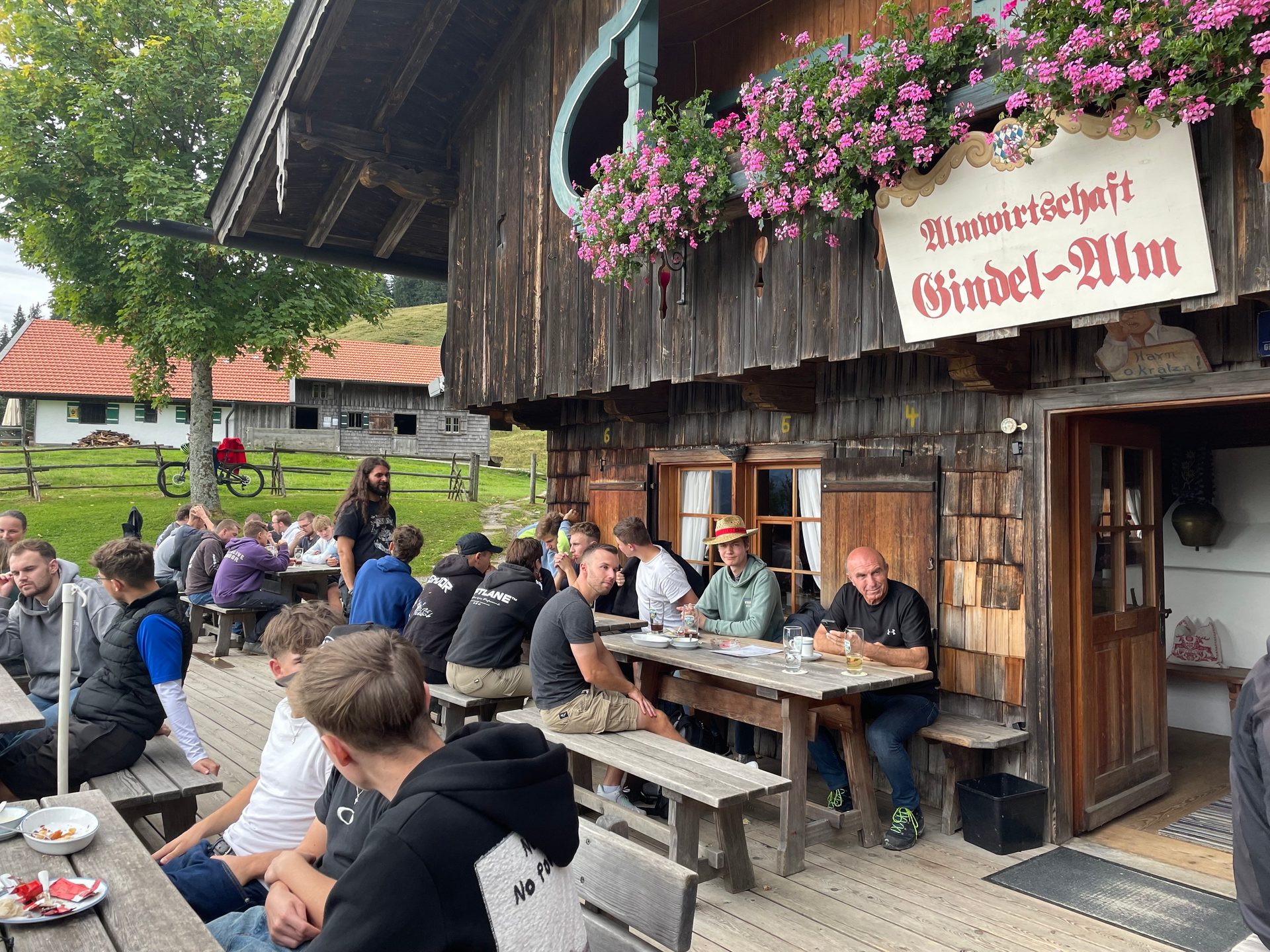 Men relaxing at wooden tables outside Gindel-Alm, a rustic mountain lodge with flowers.