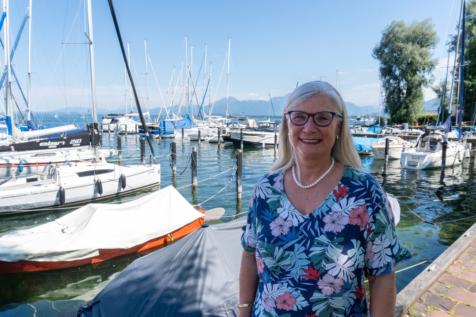 Smiling woman in glasses and a floral dress at a sunny marina with numerous sailboats and mountains.
