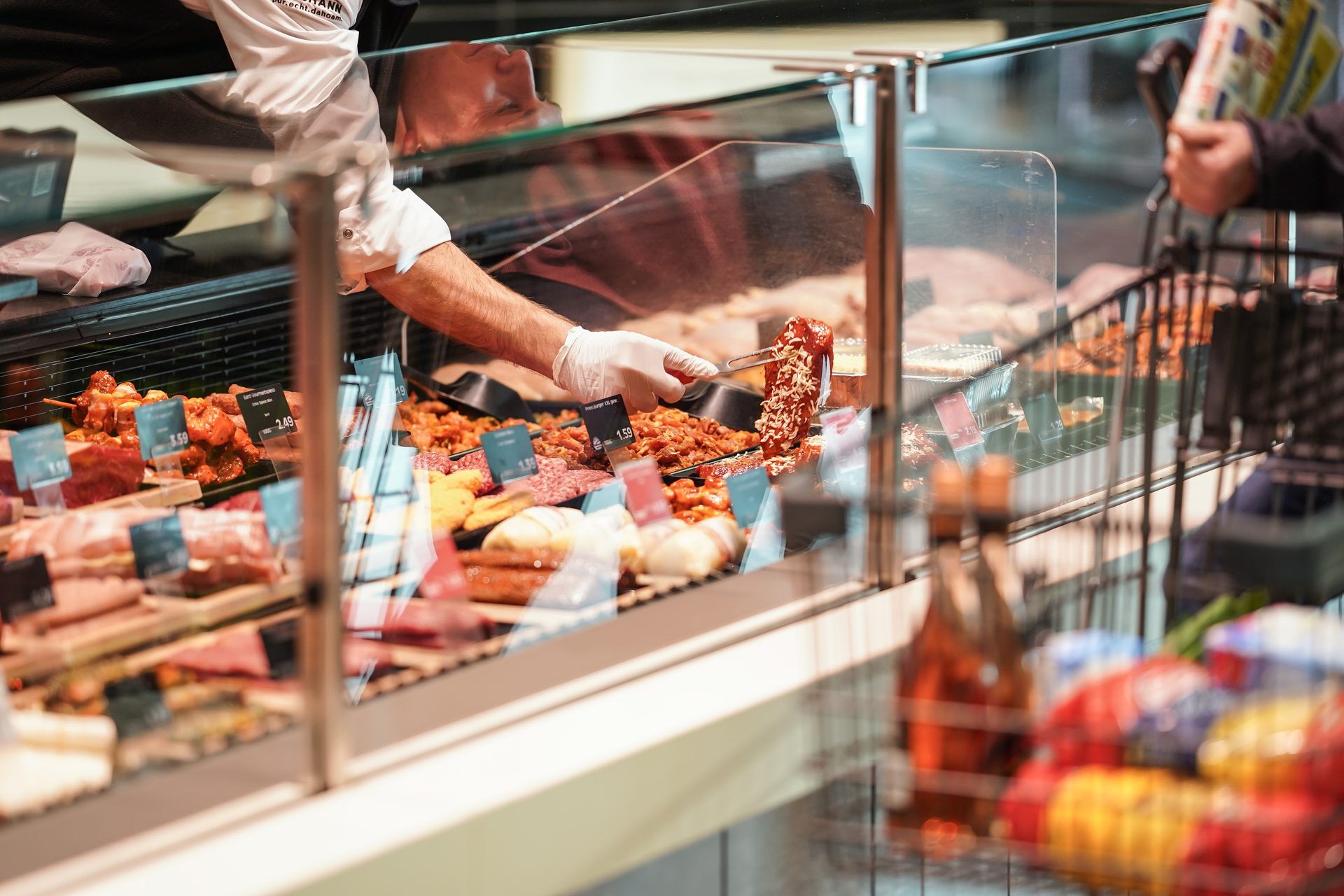 A gloved hand uses tongs to move marinated meat in a refrigerated deli case.