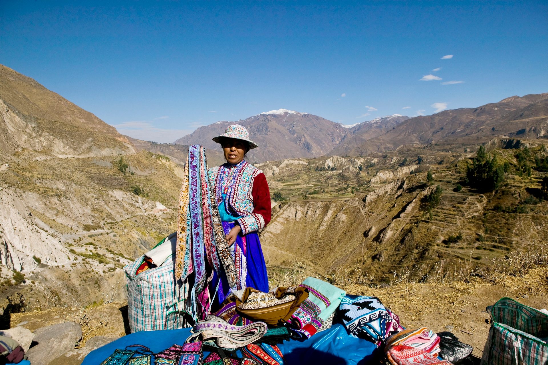 Indigenous woman in colorful traditional attire selling woven textiles in a mountainous Andean landscape.