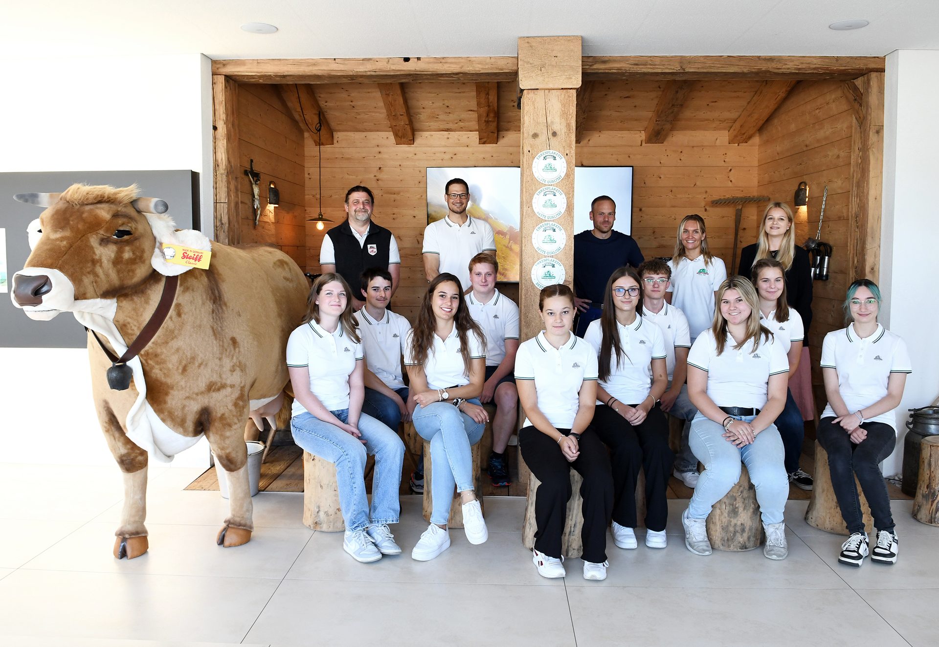 Smiling group, mostly young, with a large cow plush in a cozy, wood-paneled room.