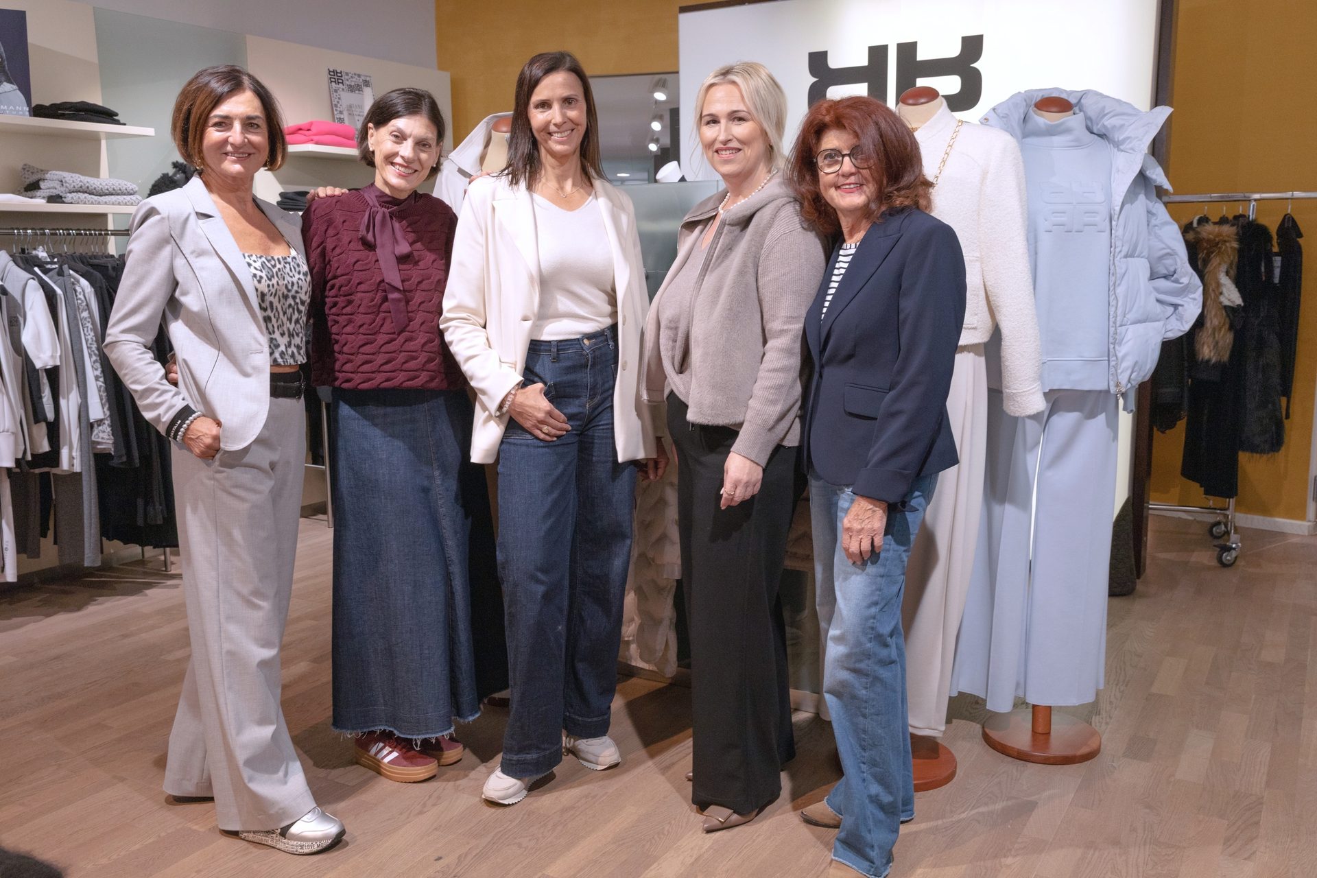 Five smiling women in chic casual outfits pose in a clothing store with mannequins and racks of clothes.