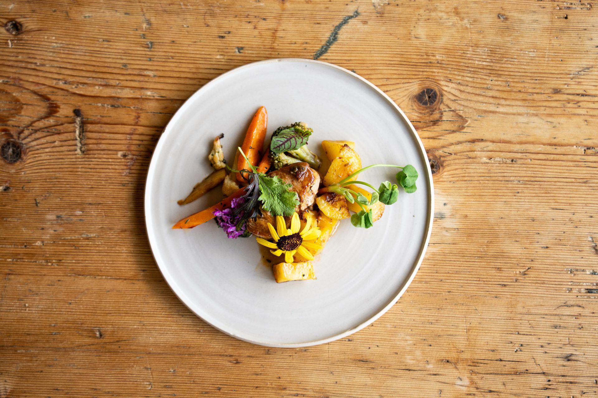 Gourmet plate with roasted vegetables, meat, and edible flowers on a white plate on a wooden table.