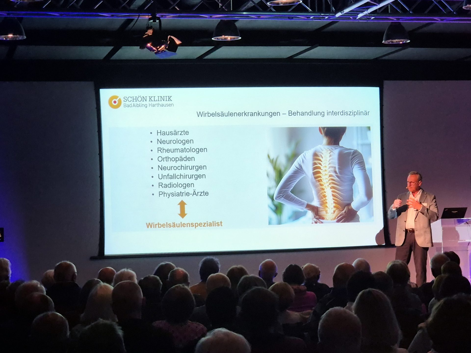 Speaker addresses audience; screen displays spine disease presentation with text and image.