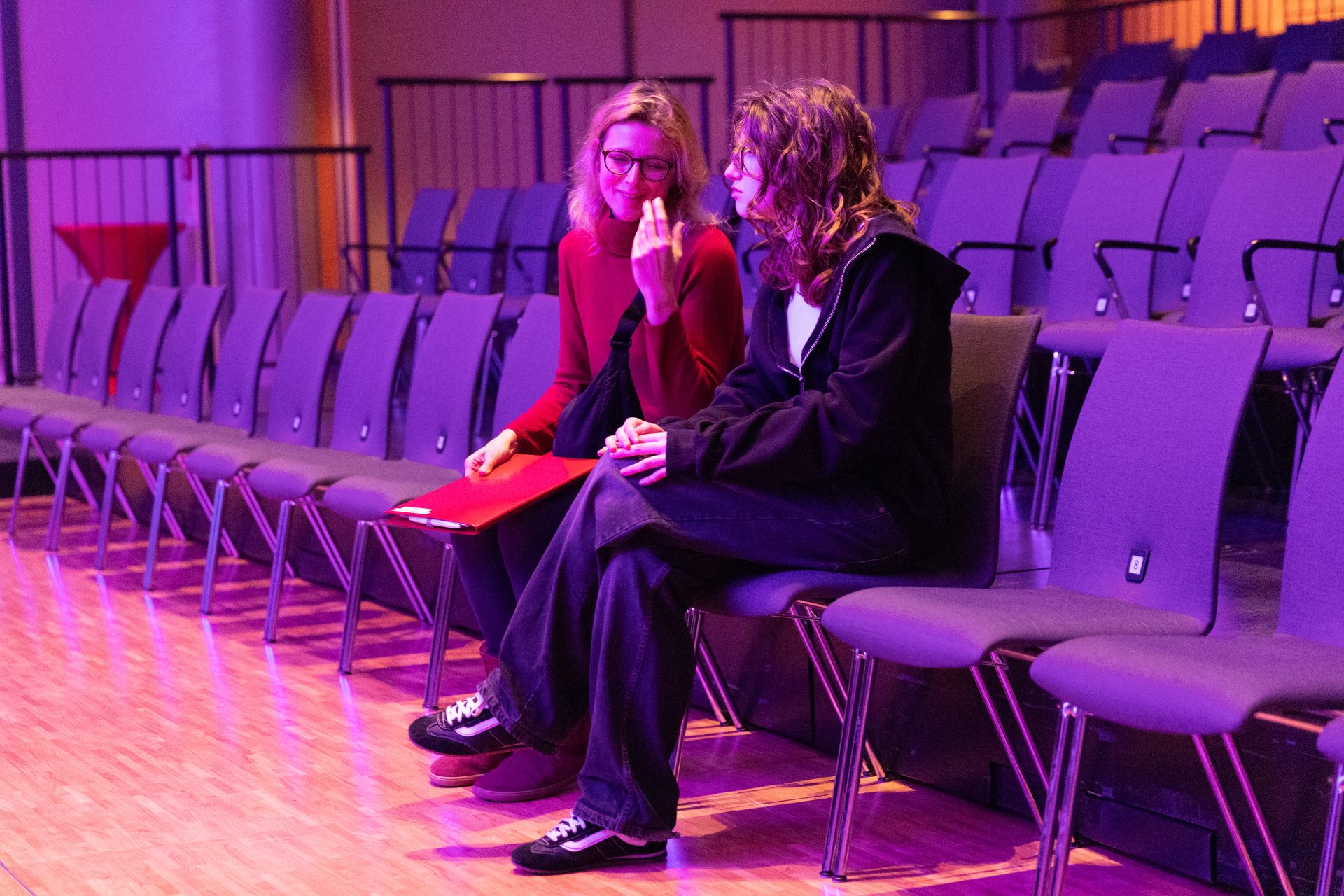 Two women in glasses talking in an auditorium under purple lights, one holding a red folder.
