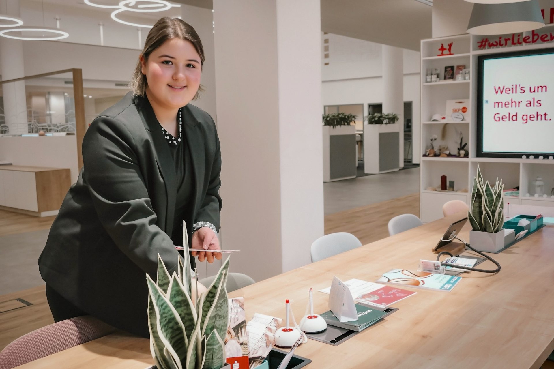 Smiling woman in a modern bank, leaning over a table with plants and documents.