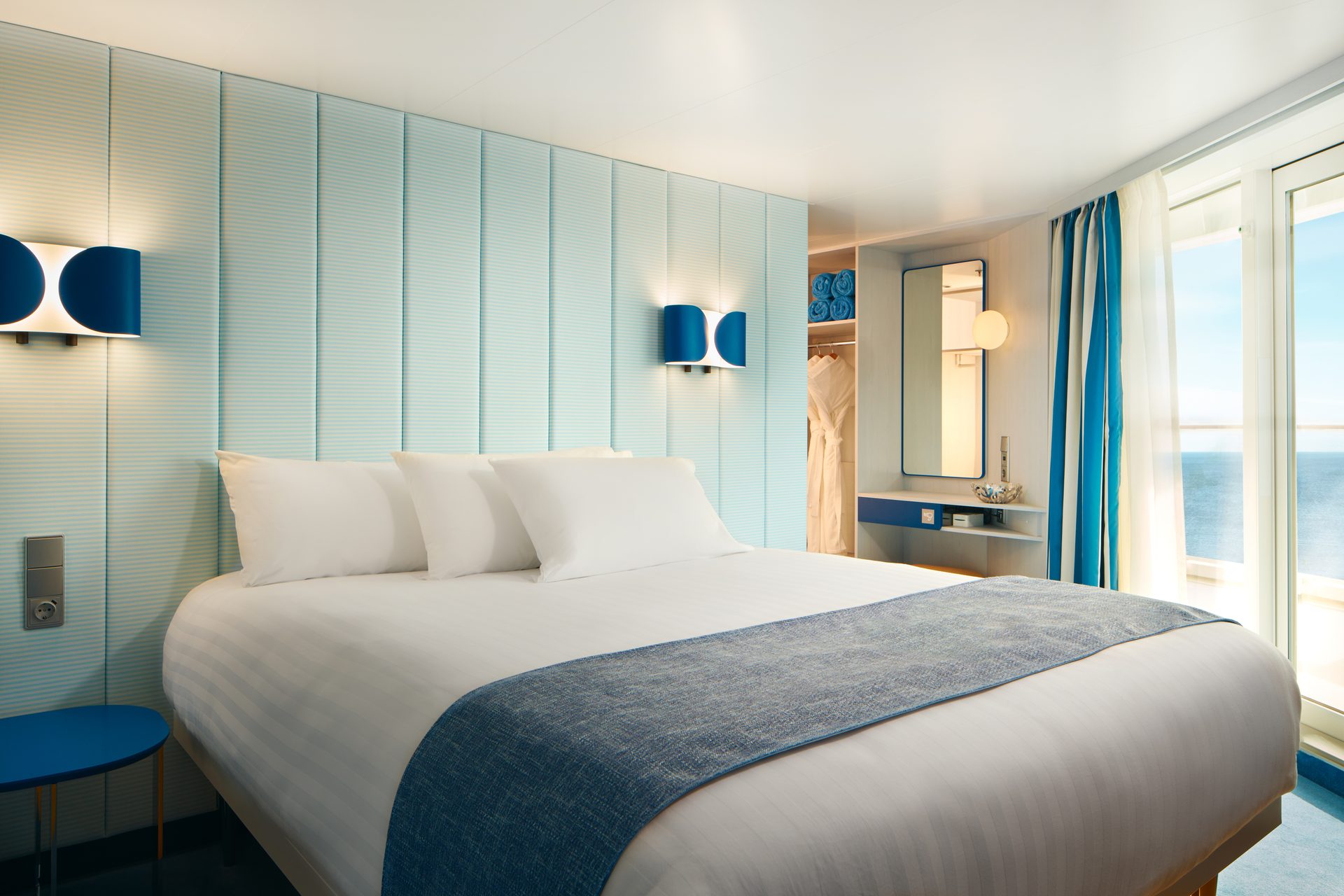 Modern cruise cabin with a large bed, blue and white decor, and an ocean view from the window.