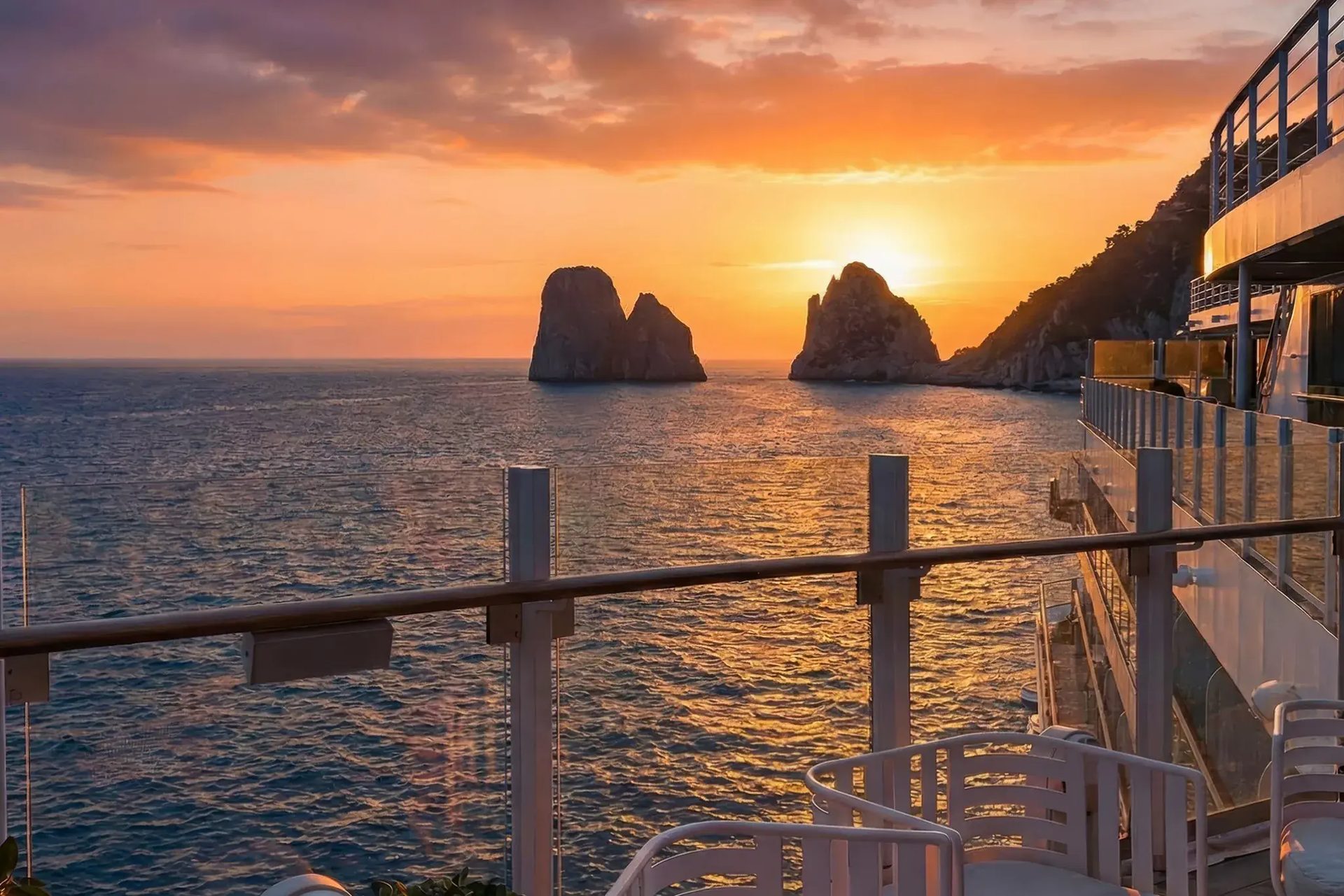A vibrant sunset over the sea, featuring prominent sea stacks, viewed from a cruise ship deck.