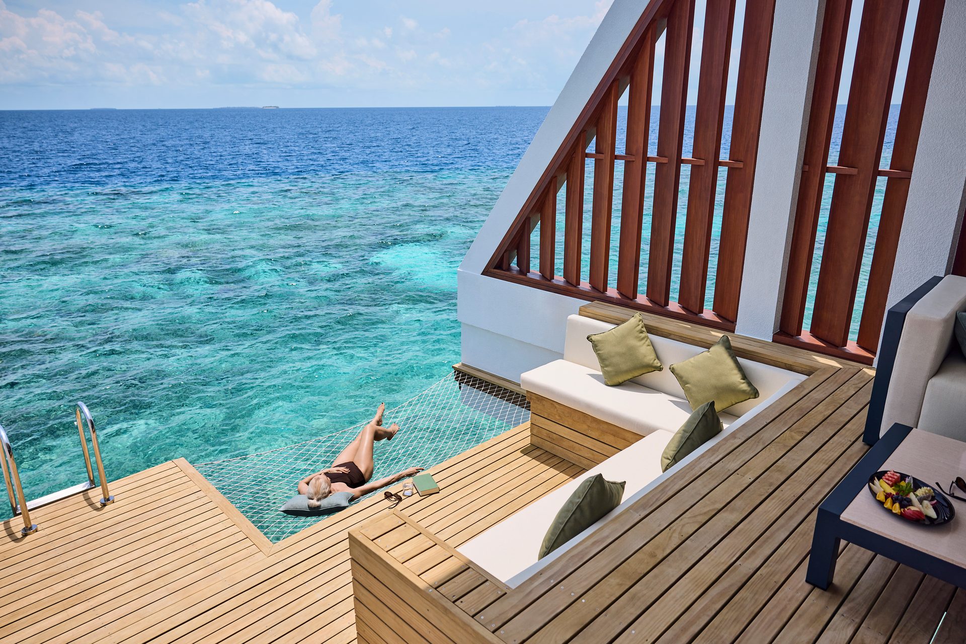 Woman on hammock over clear ocean next to a wooden deck with lounge seating.