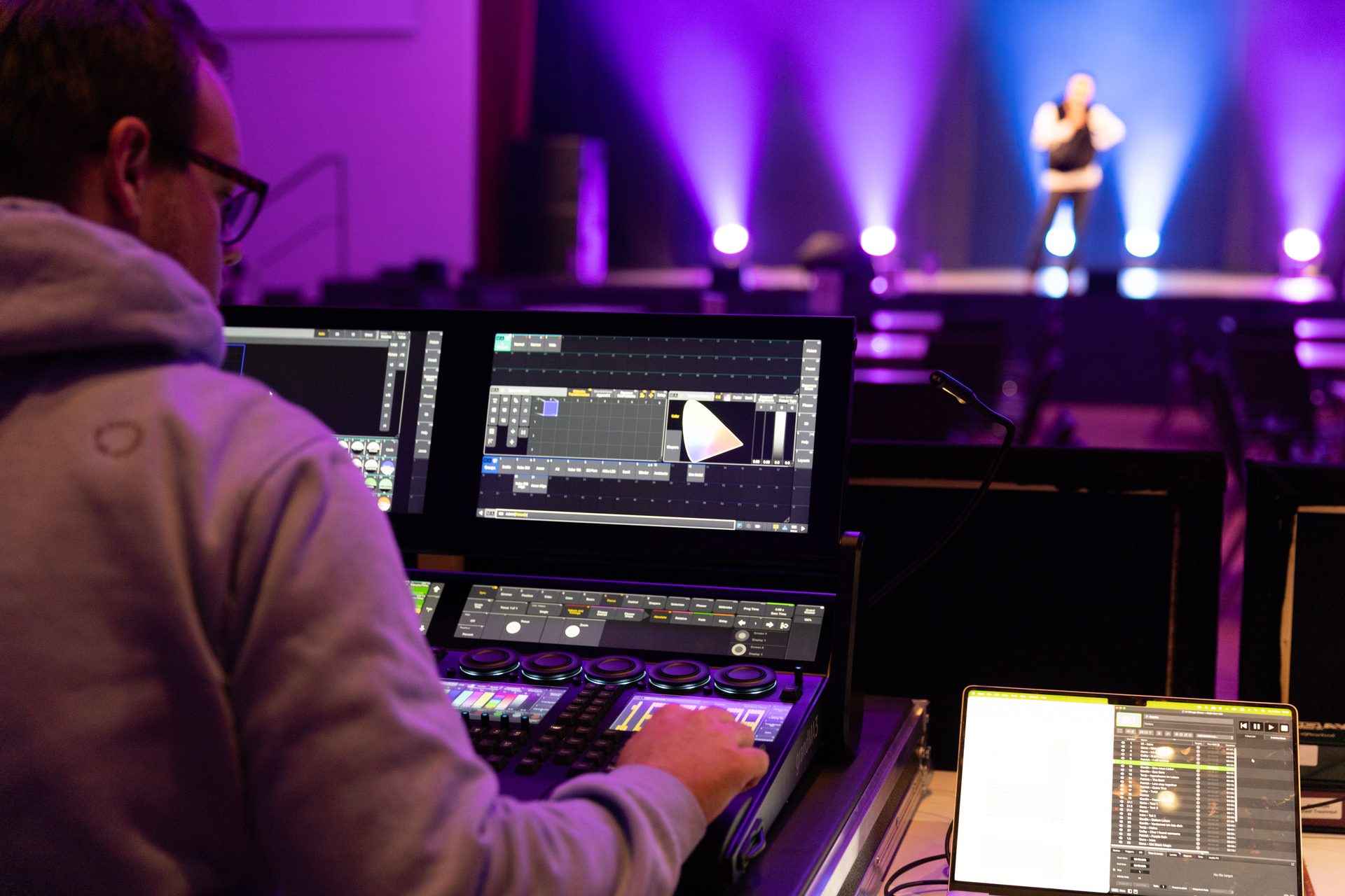 Man operates a lighting console for a performer on a stage with purple lights.