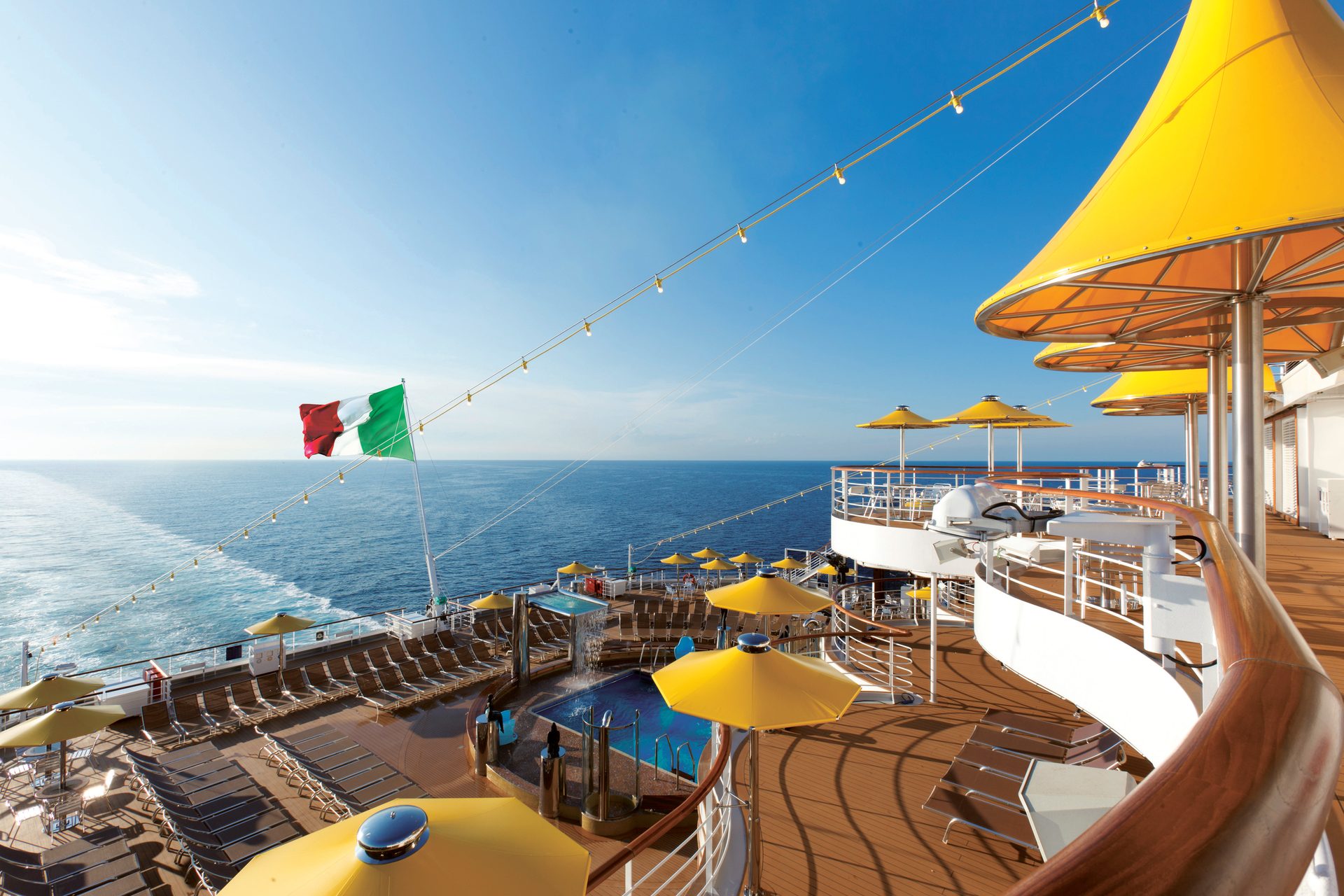 Deck of a cruise ship with an Italian flag, yellow umbrellas, a pool, and ocean view with a wake.