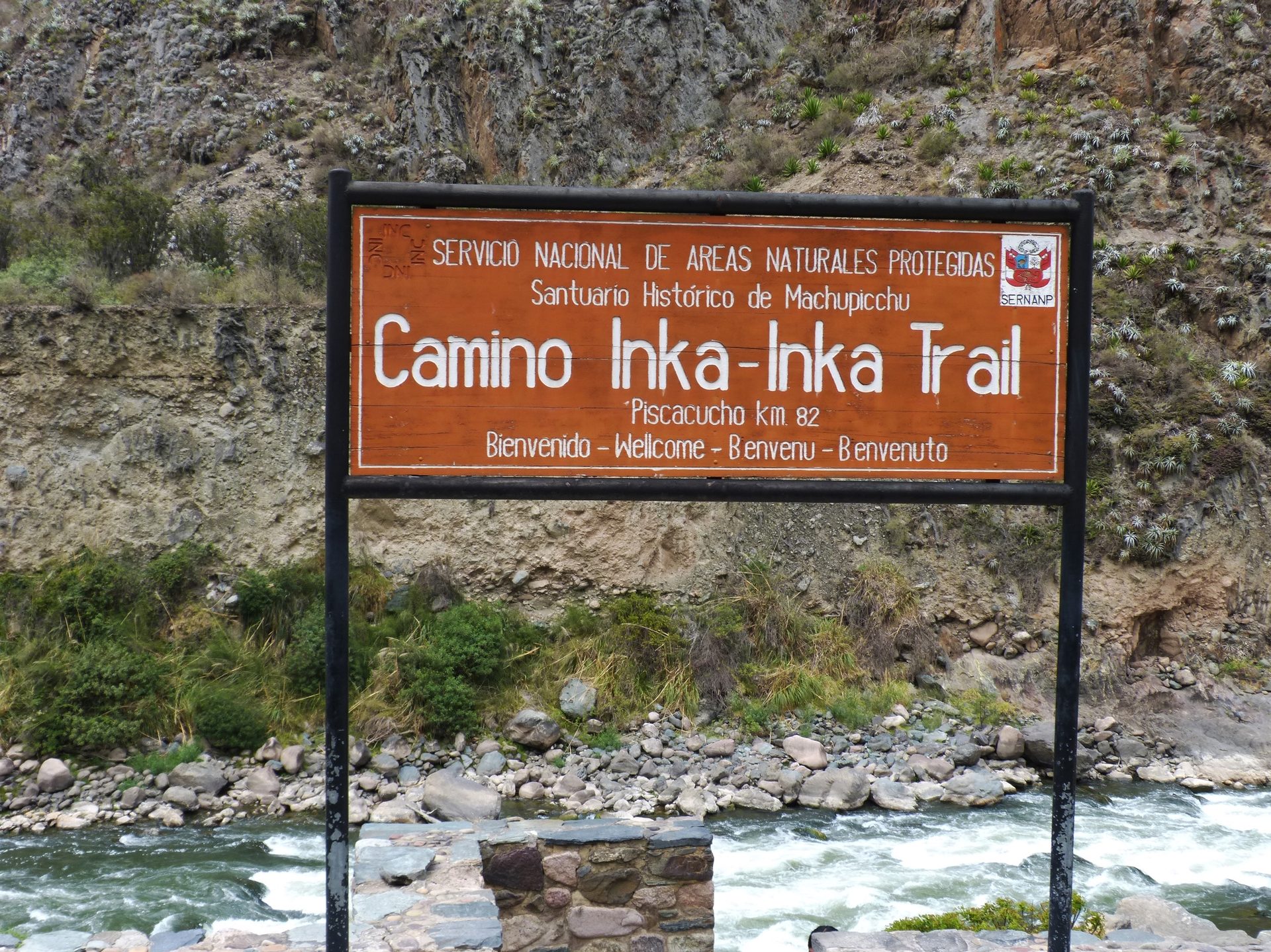A wooden sign marks the start of the Inca Trail to Machu Picchu, with a river and mountains in the background.